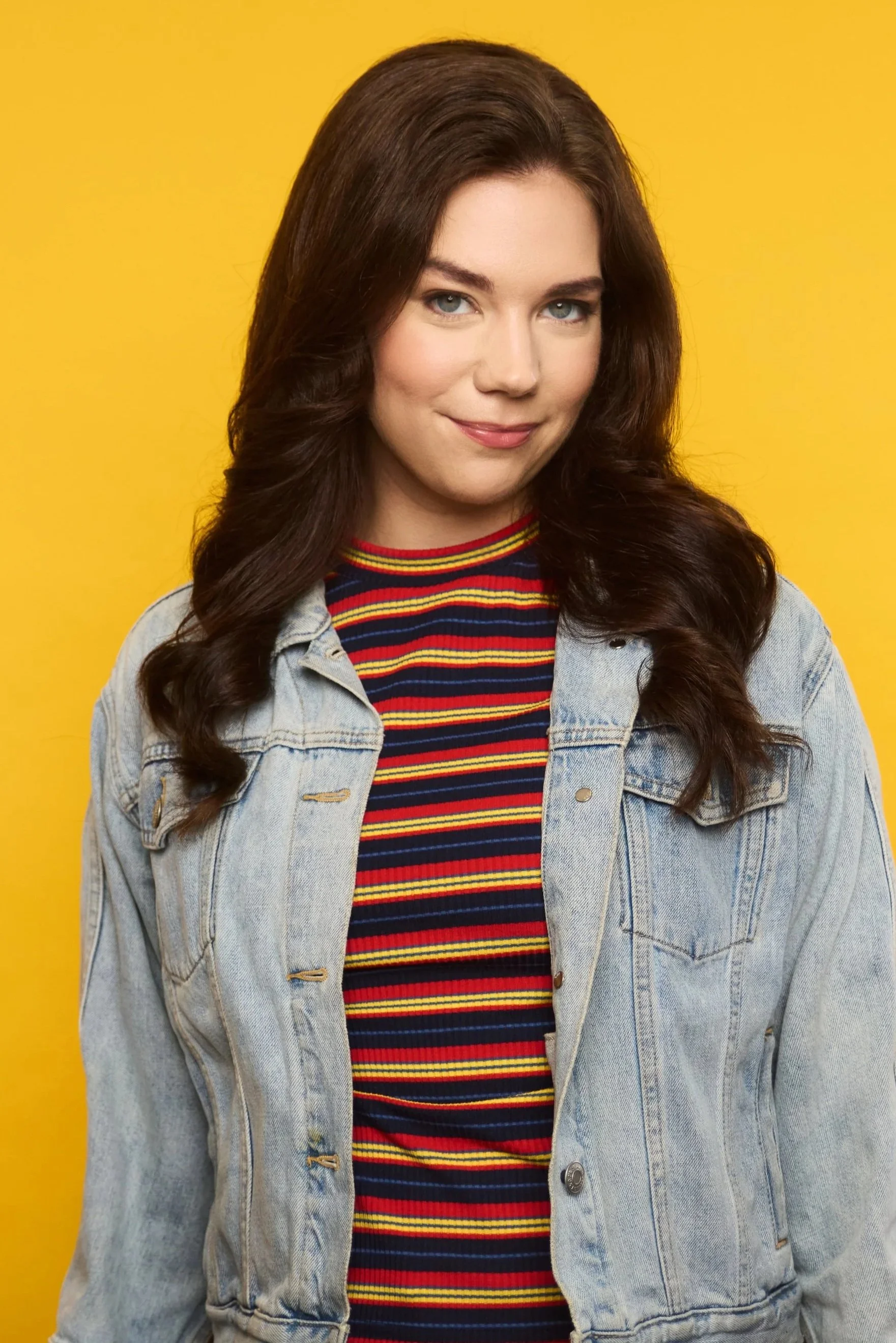 A young woman with long, wavy brown hair and blue eyes wearing a multicolored striped shirt and a light denim jacket against a yellow background.