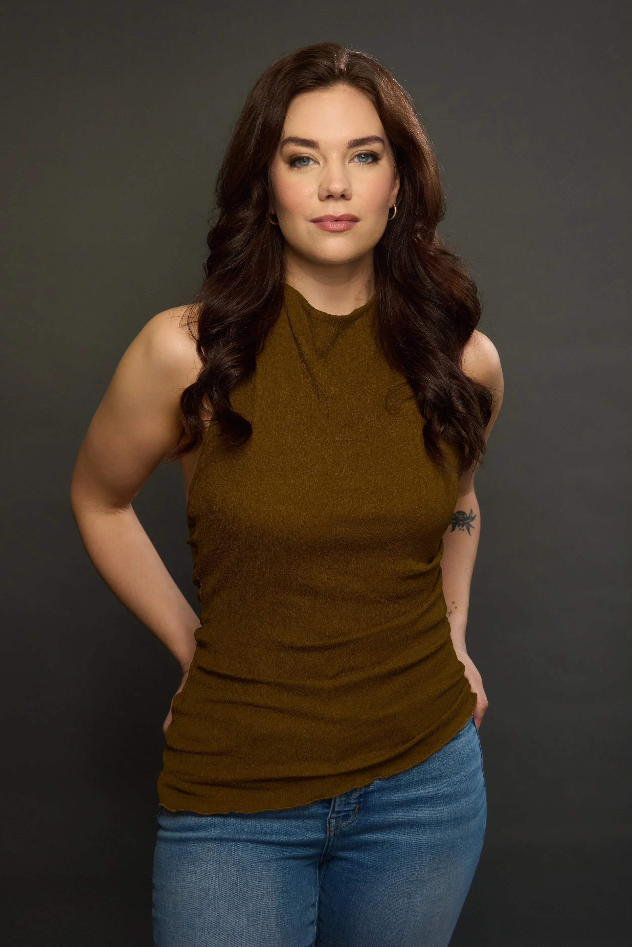 Portrait of a young woman with long wavy brown hair, wearing a sleeveless olive-green top and blue jeans, standing against a dark gray background.
