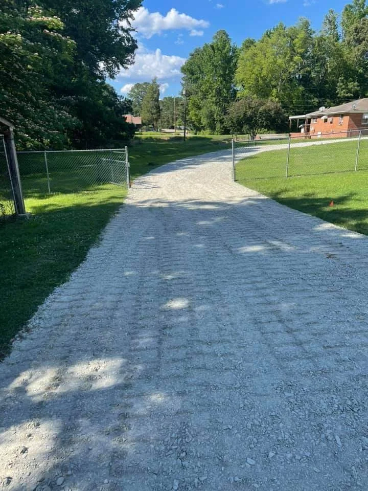 Gravel driveway leading to a residential neighborhood, fenced yard on both sides, with trees and houses in the background under a partly cloudy sky.