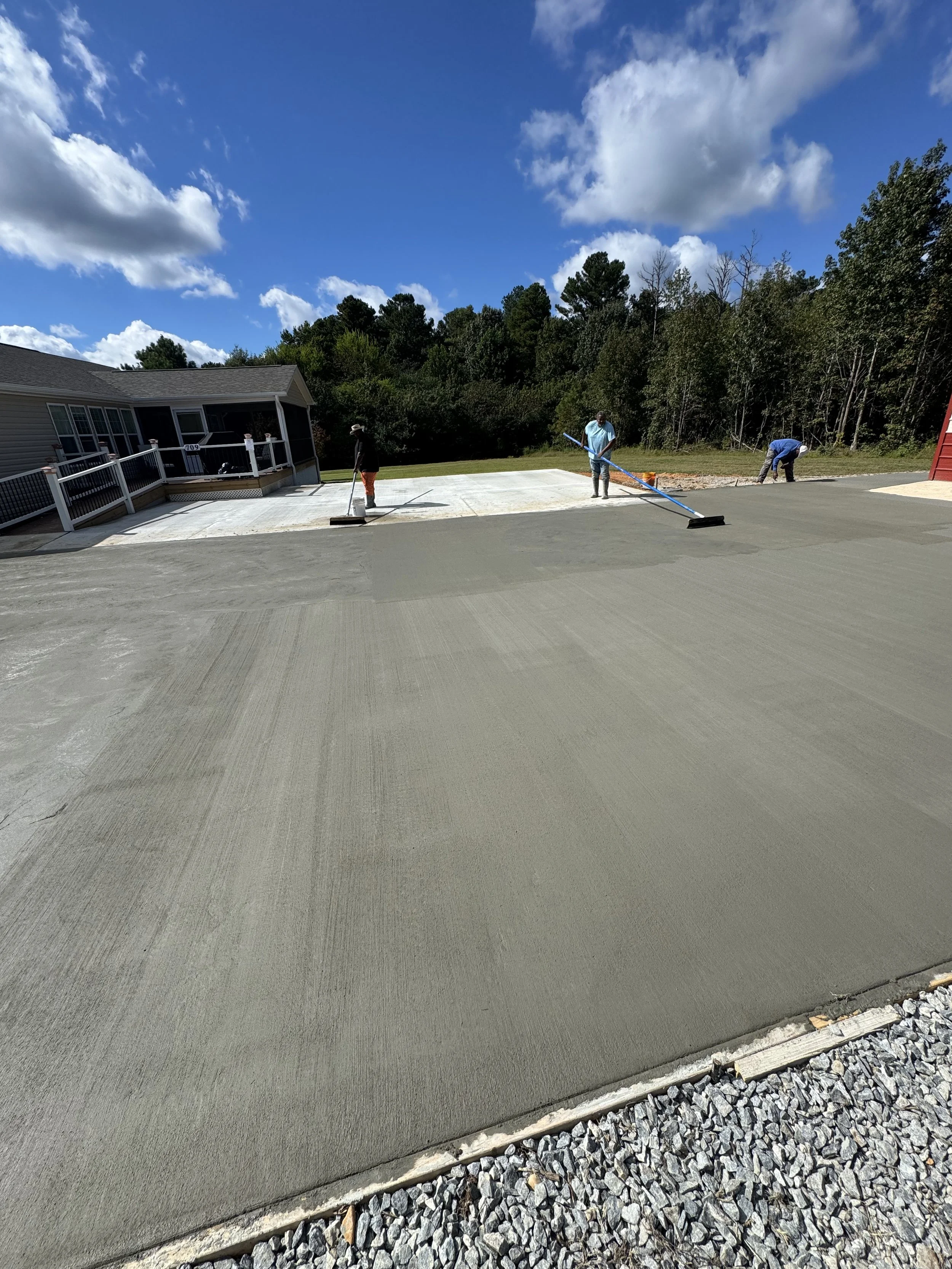 Three workers are finishing a concrete driveway, with one using a long pole to smooth the surface, on a bright sunny day with scattered clouds and a woods background.