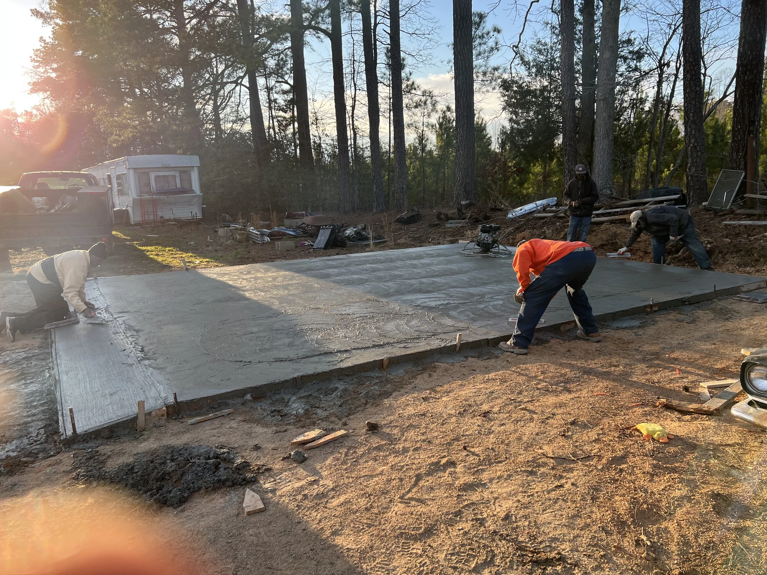 Construction workers leveling and smoothing wet concrete on a large slab outdoors in a wooded area during late afternoon or early evening sunlight.