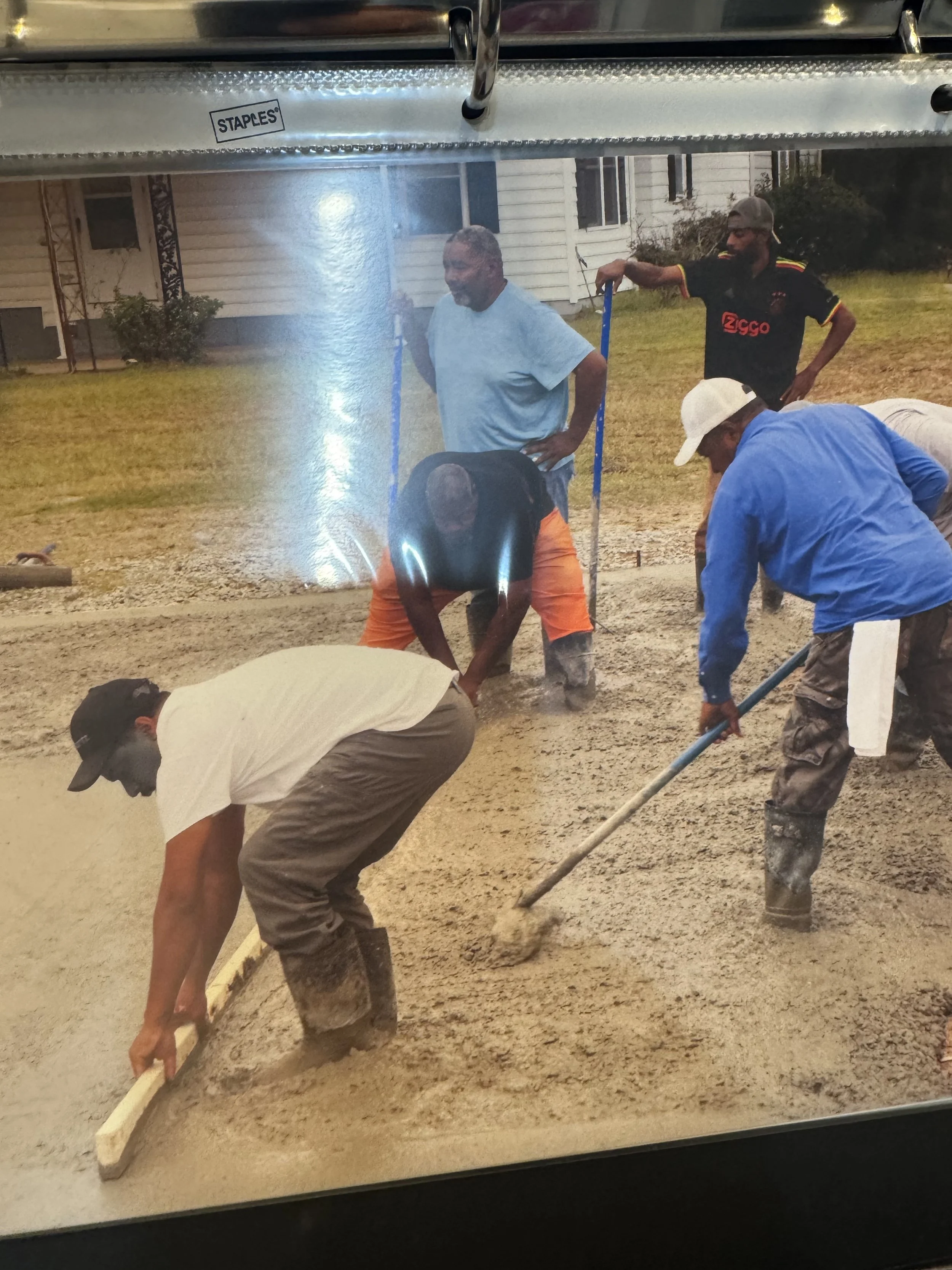 People working together to pour and level concrete on a slab, some using shovels and trowels, outdoors in a residential area.