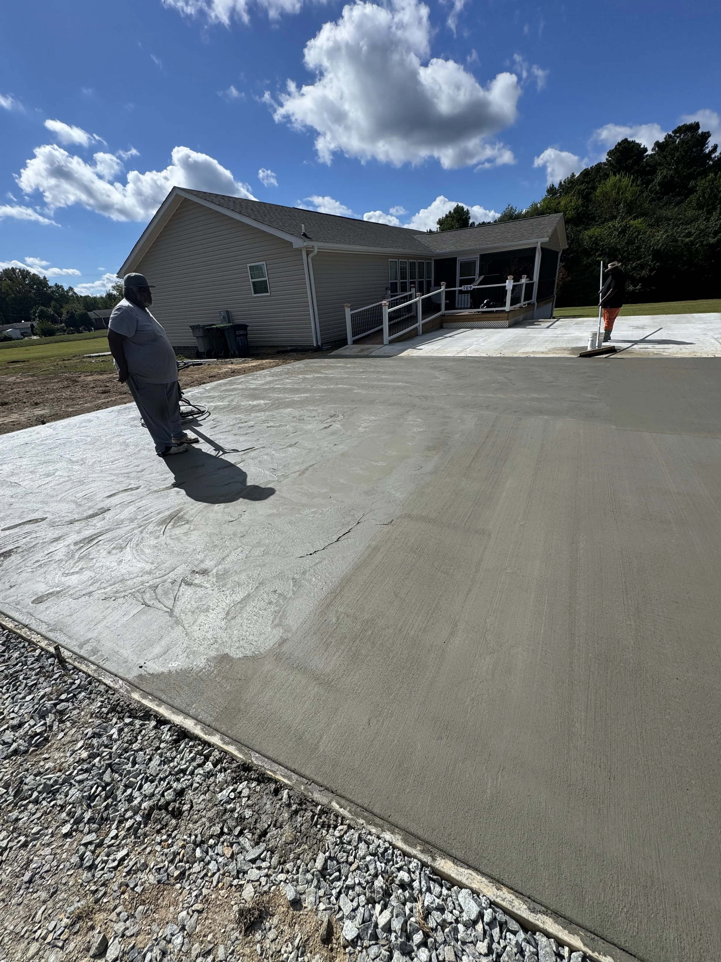 Two men are applying freshly poured concrete to a driveway in front of a house, with the sky partly cloudy overhead.