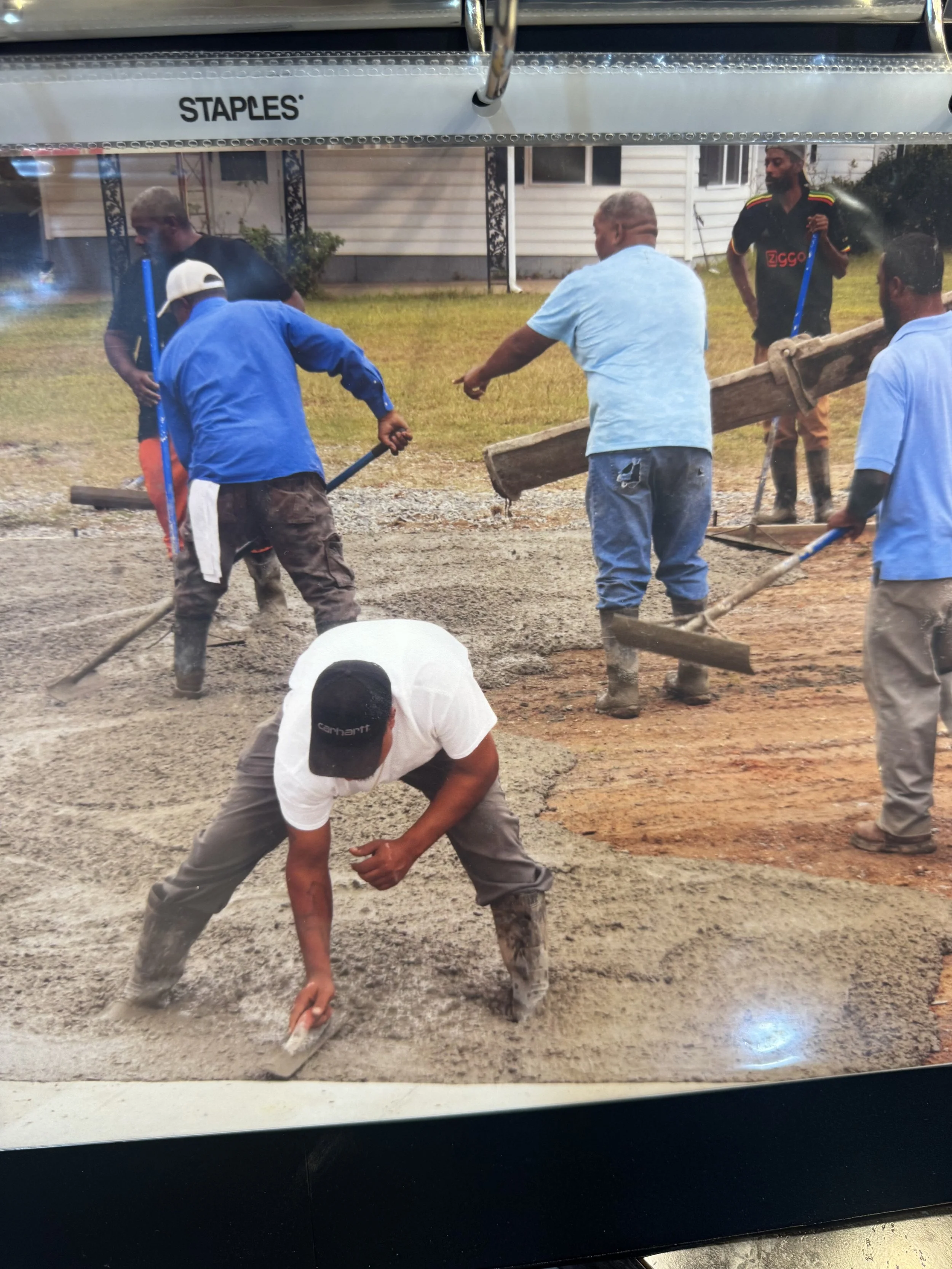 Group of men working together pouring and smoothing concrete on a driveway or pathway, with some men using shovels and a worker in the foreground spreading concrete.