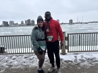 Two people in athletic clothing standing together on a snowy waterfront with a city skyline in the background.