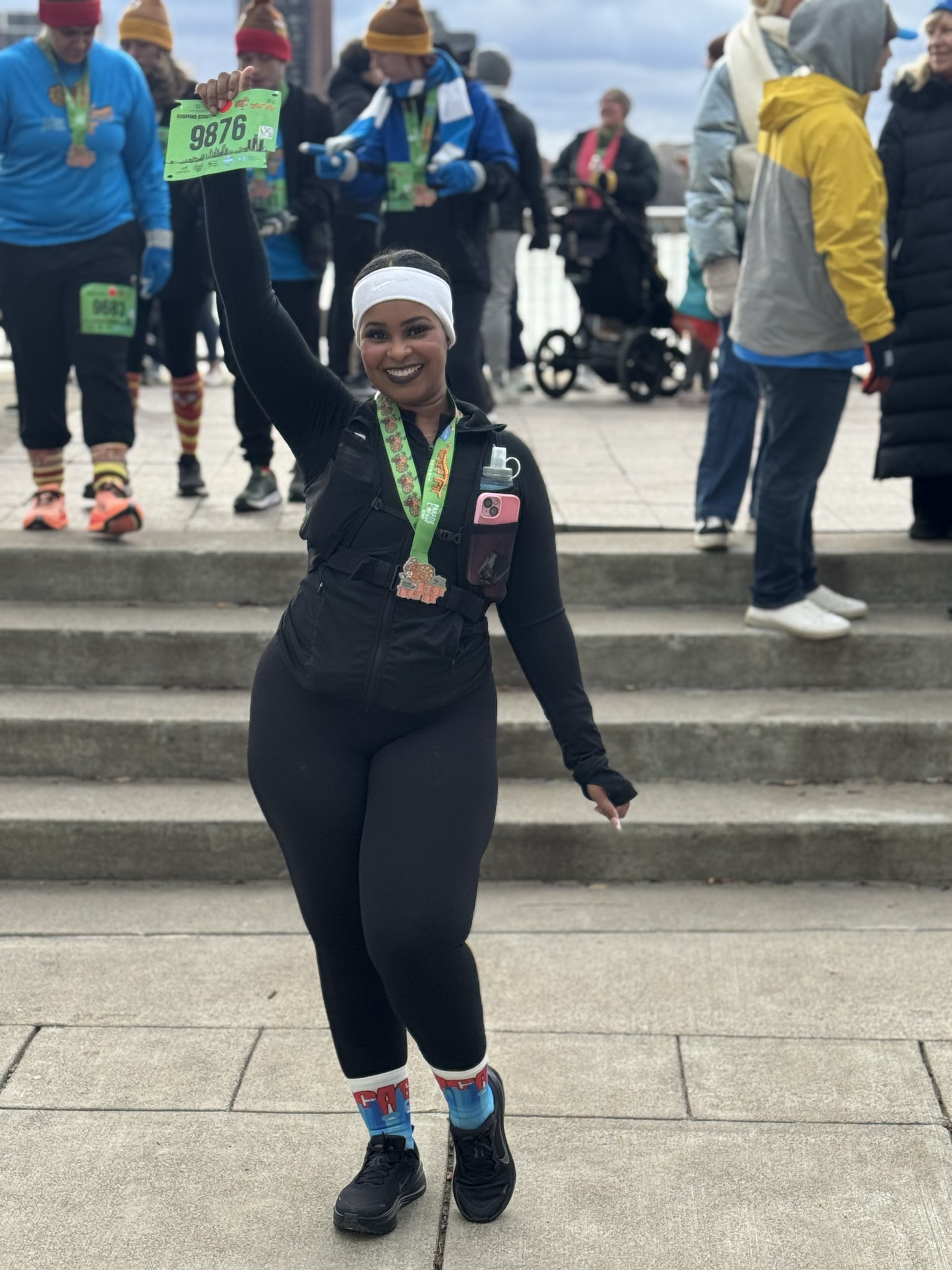 A woman happily holding a green race bib with the number 9876 after completing a charity run. She is dressed in black athletic wear, wearing a white headband, and has a medal around her neck. She is smiling and standing in front of a group of runners and supporters on the steps outdoors.
