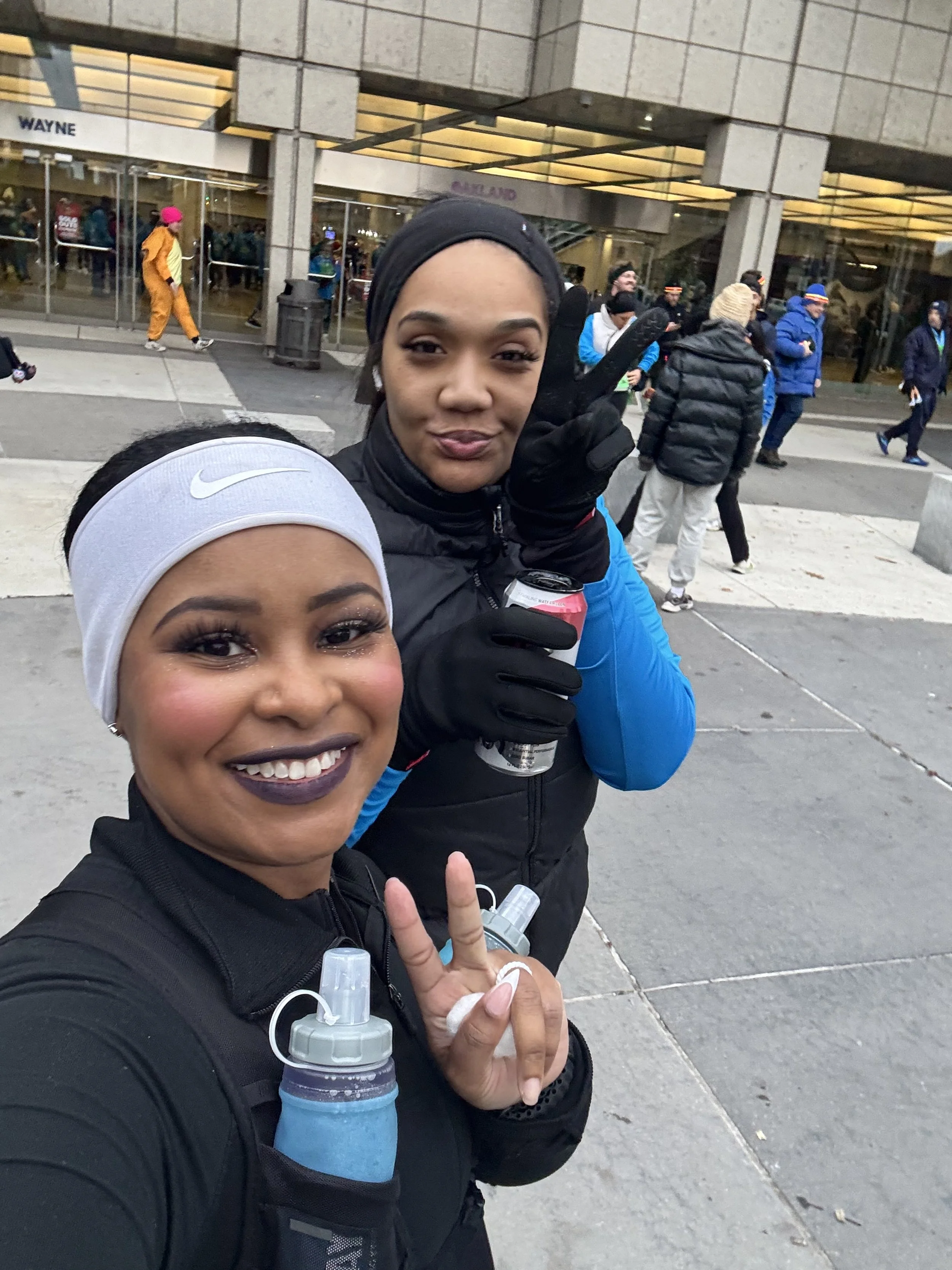 Two women smiling and taking a selfie outside a building with a crowd in the background. One woman is wearing a white headband, black athletic clothing, and has a water bottle in her gear. The other woman is making a peace sign and holding a beverage, wearing black gloves and a black and blue athletic jacket.