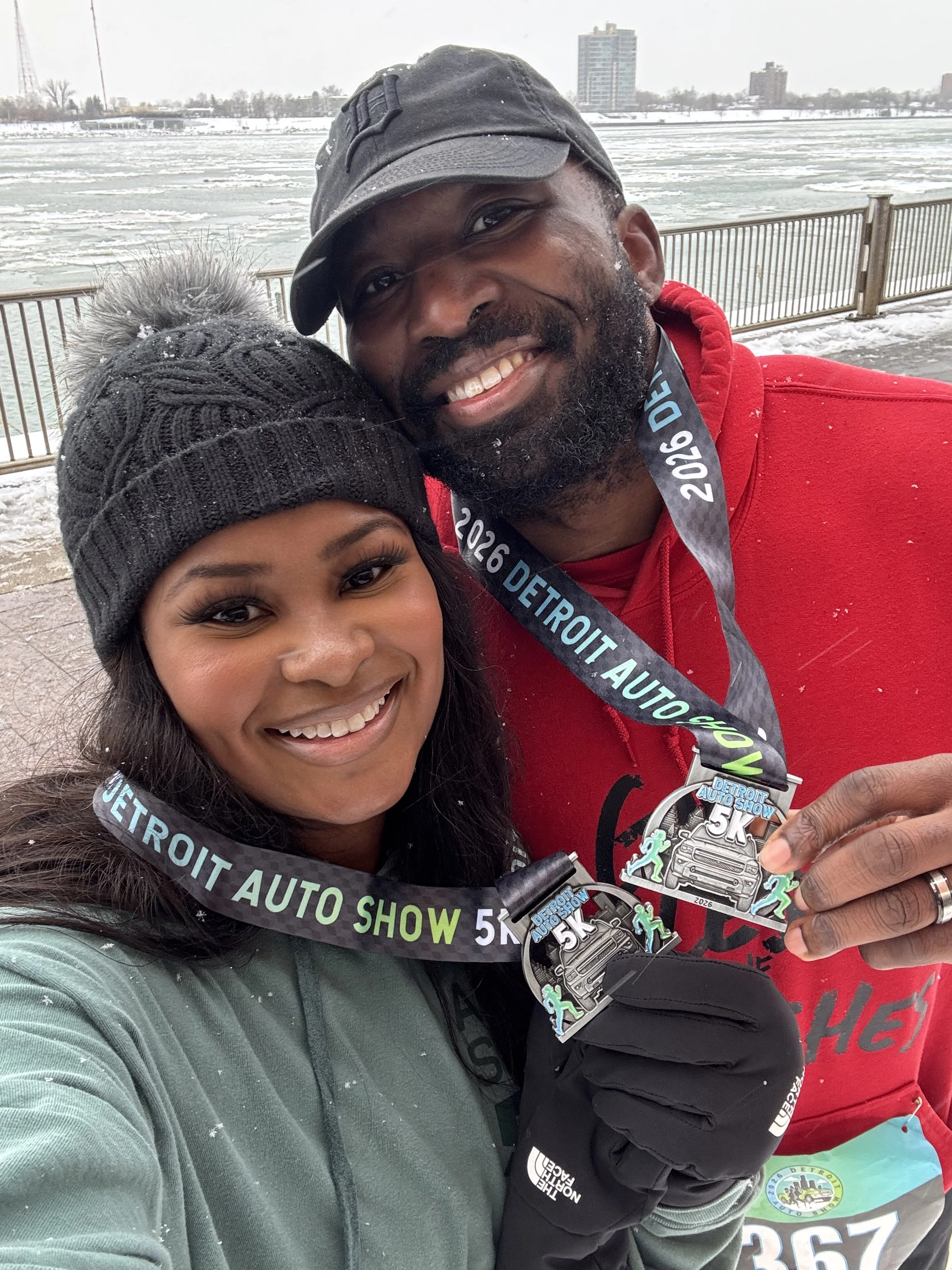 Two people smiling with medals at the Detroit Auto Show 5K in winter, standing outdoors near a frozen river, both dressed in warm clothing.