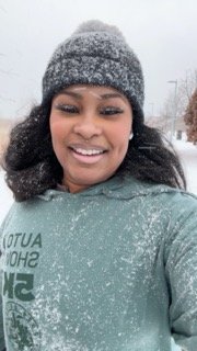 Woman smiling outdoors in winter weather, wearing a gray beanie and a greenish-gray hoodie, with snow falling around her.