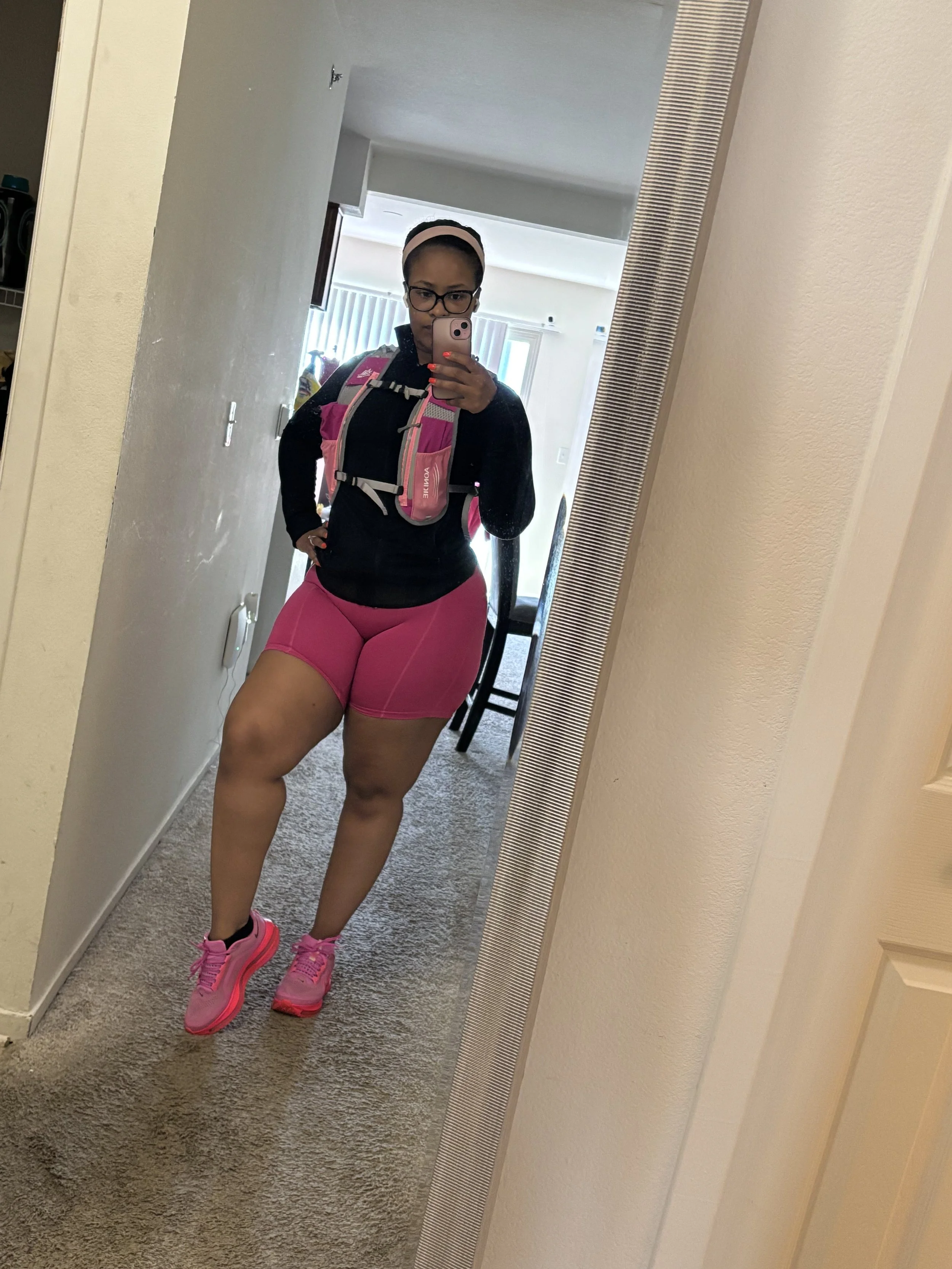 A woman in athletic outfit taking a mirror selfie, wearing pink shorts, pink running shoes, black long-sleeve top, glasses, and a pink backpack.