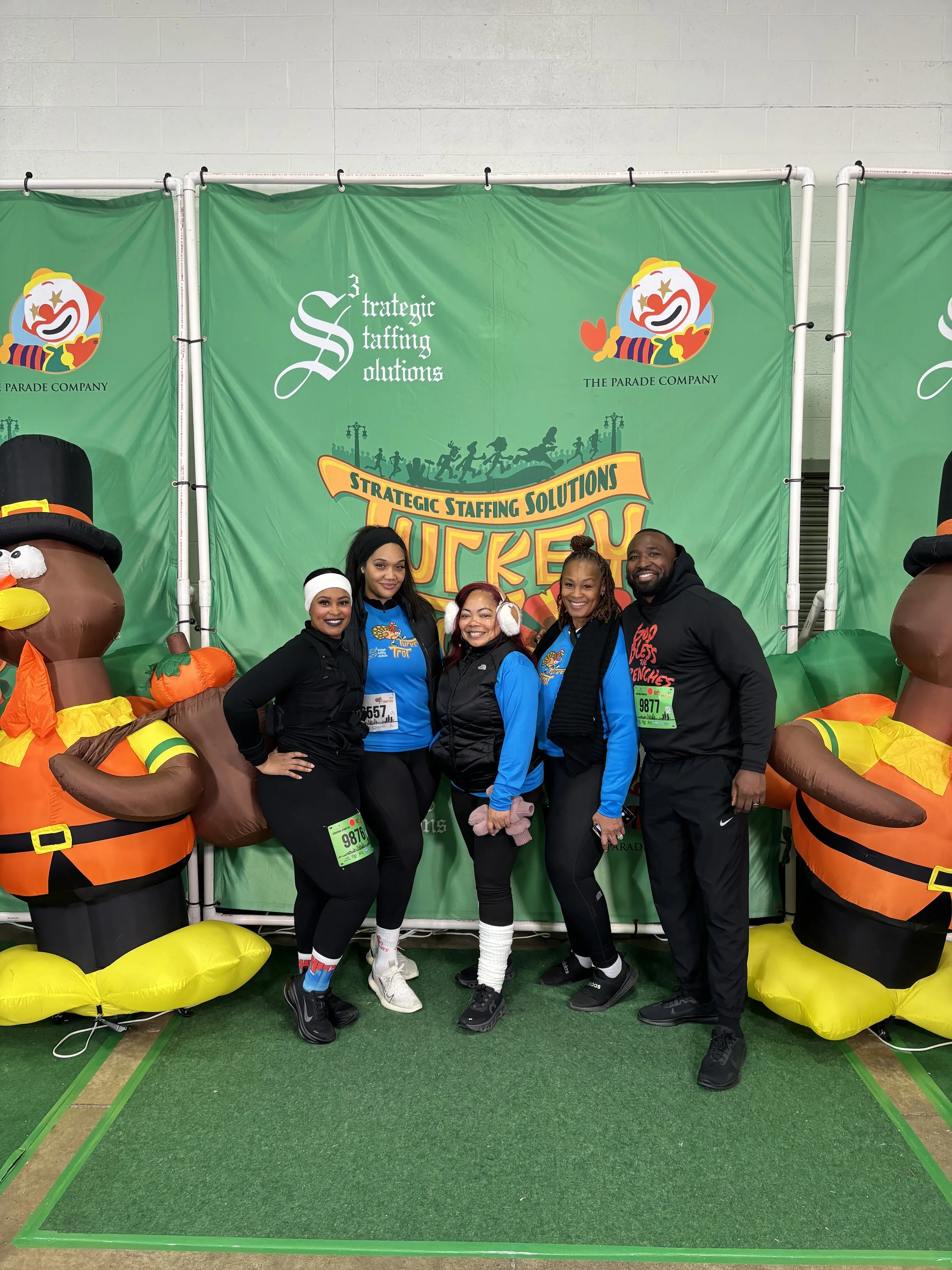 Group of five people posing in front of a green backdrop with Christmas-themed Inflatable turkey decorations on either side, and balloons at the top, at a festive event.