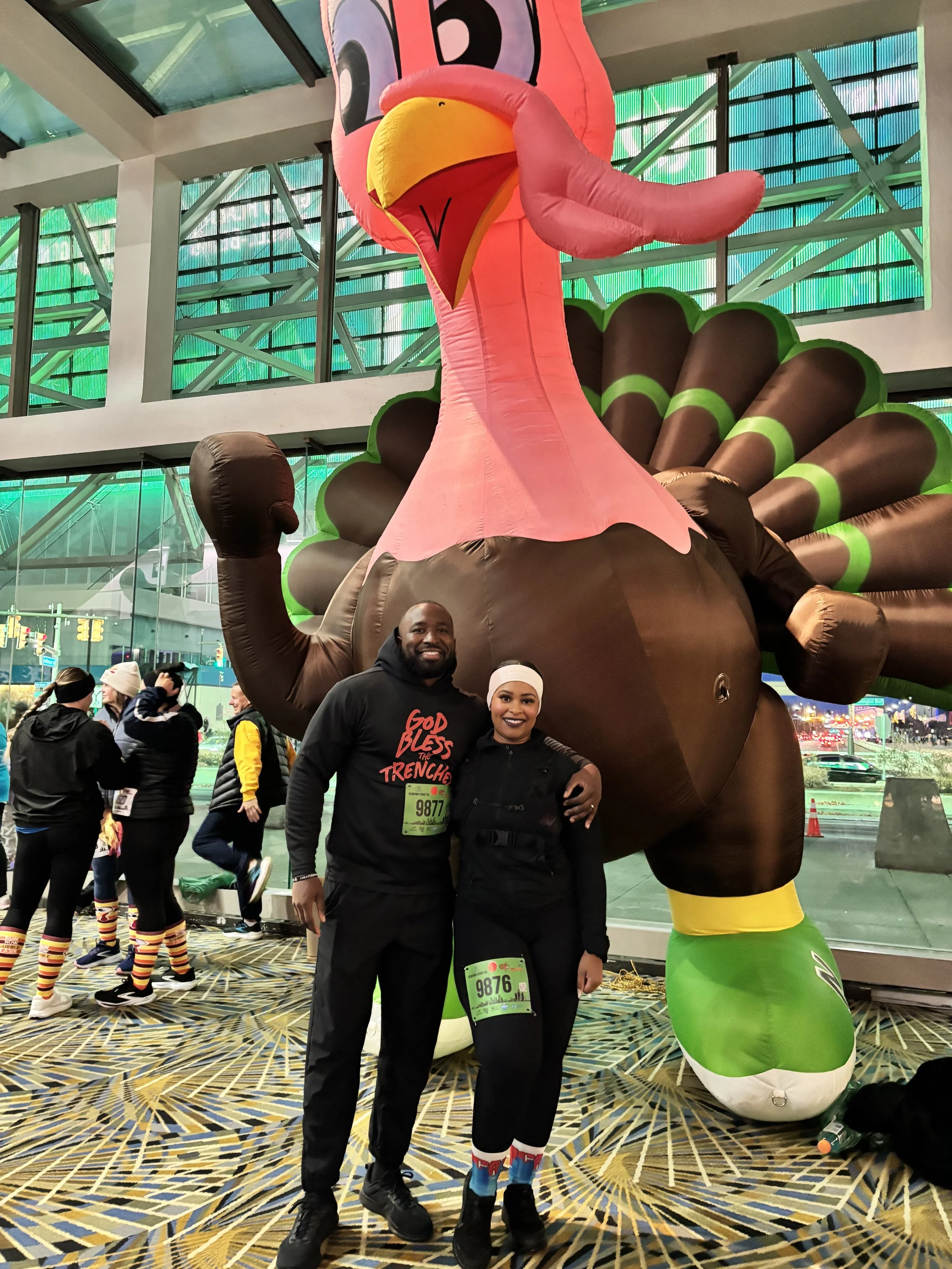 Two people smiling and posing in front of a large inflatable turkey mascot at a festive indoor event, with other participants in the background.