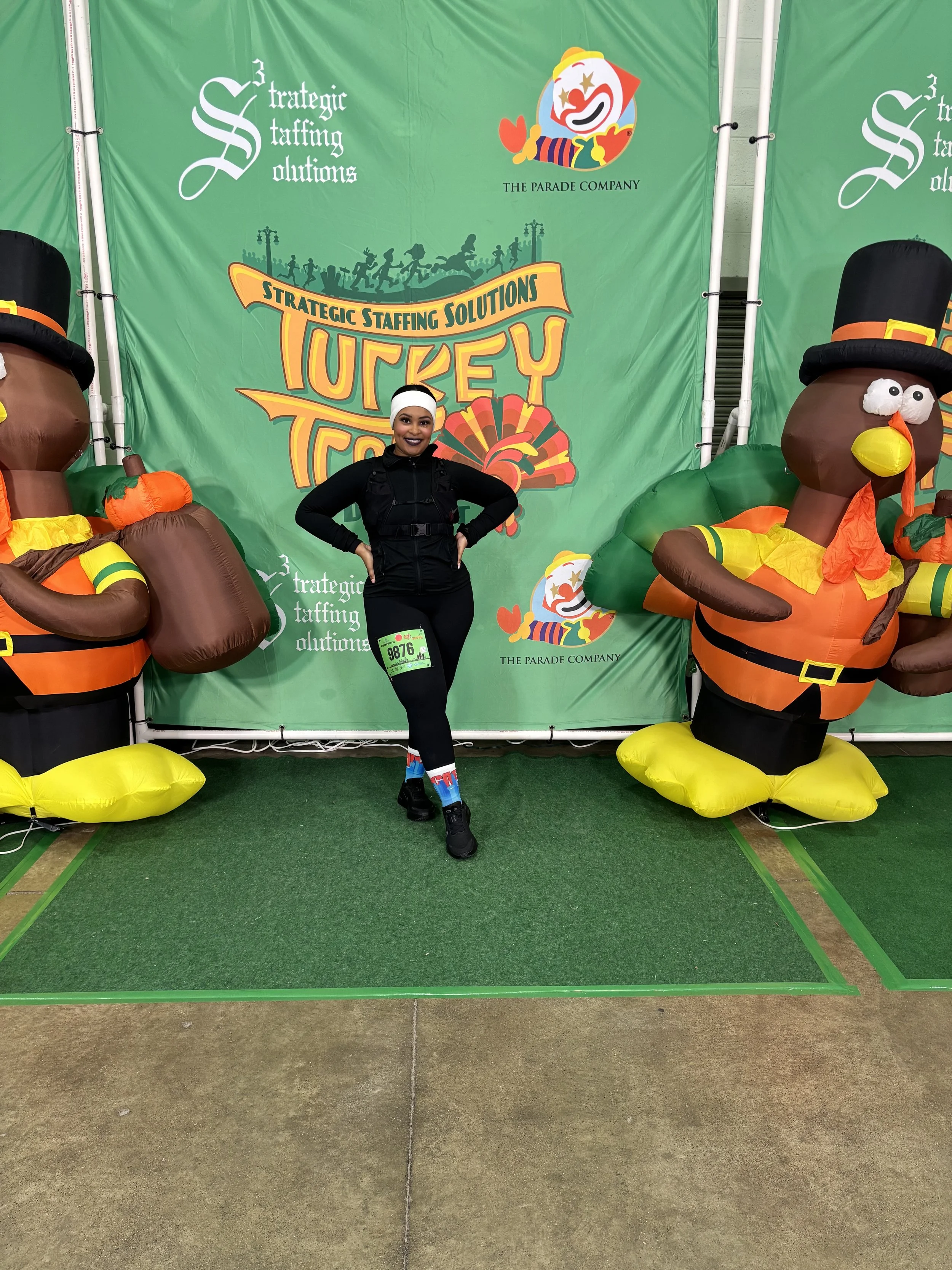 Woman standing between two large inflatable turkey decorations, smiling, at an event with a green backdrop displaying the event name 'Turkey Trot' and logos for 'Strategic Staffing Solutions' and 'The Parade Company.'