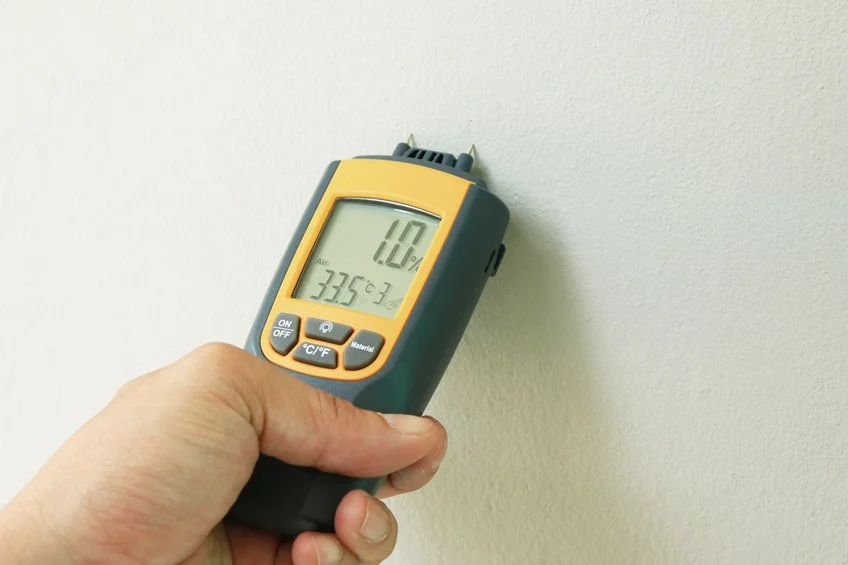 Person holding a digital infrared thermometer against a beige wall, with the display showing a temperature of 33.5 degrees Celsius.