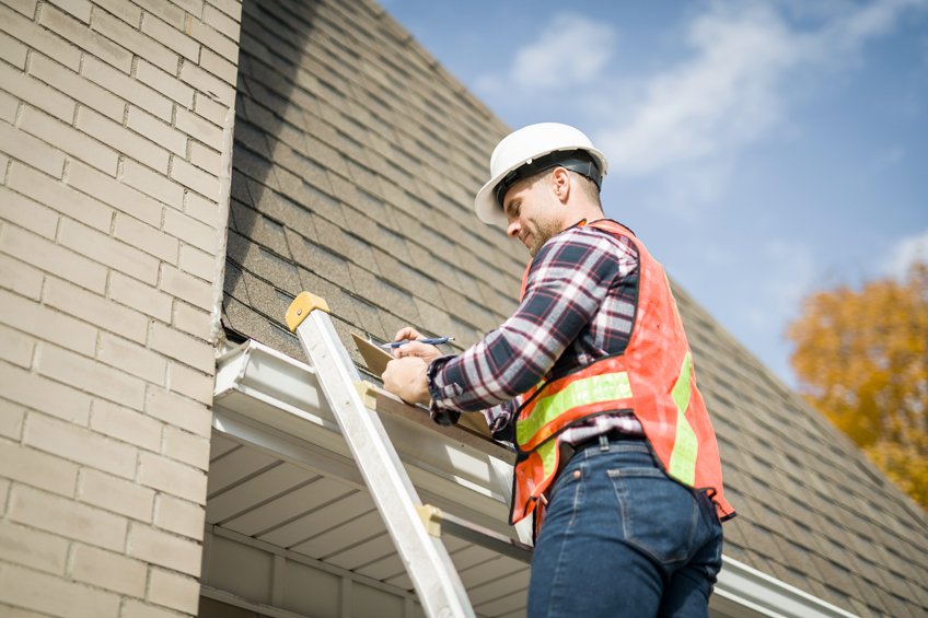 A man wearing a white safety helmet, plaid shirt, and orange safety vest standing on a ladder, working on the roof gutter of a house against a background of blue sky and autumn trees.