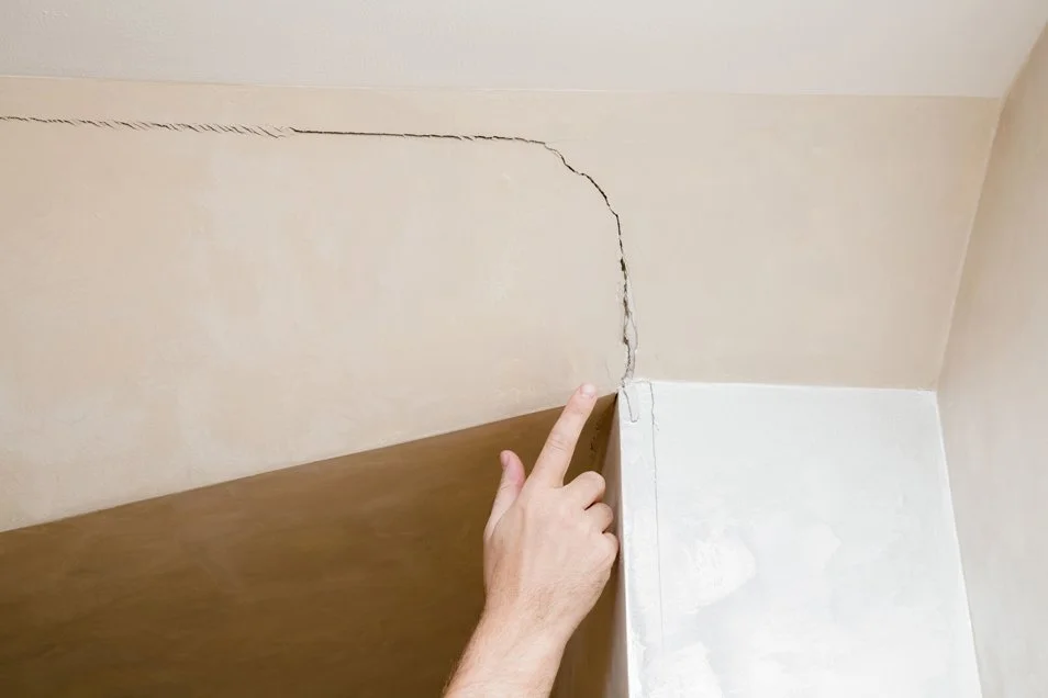 A person's hand pointing at a crack in the wall near the ceiling of a room.