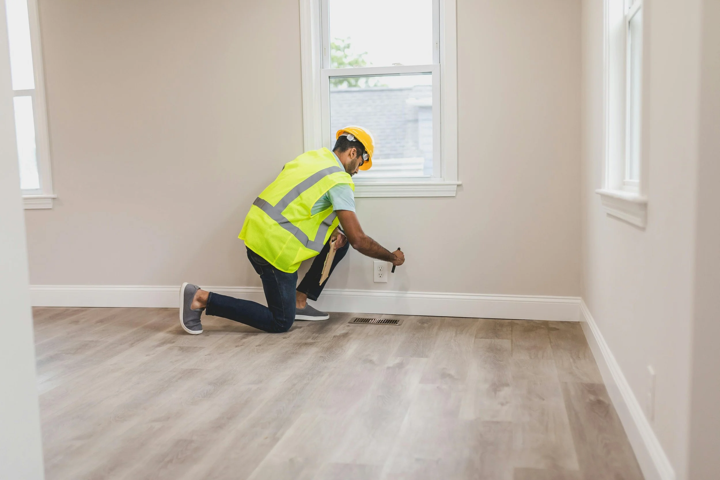 A man in a yellow safety vest and hard hat kneeling on the floor of a room, working near an electrical outlet.