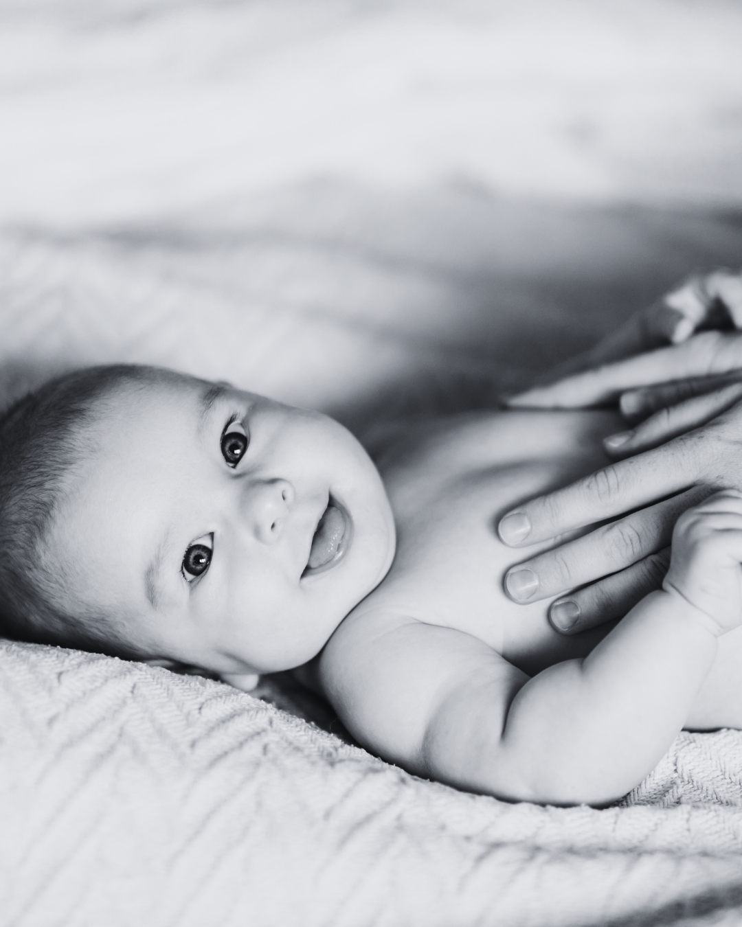 Black and white photo of a smiling baby lying on a textured surface, with adult hand gently touching their chest.