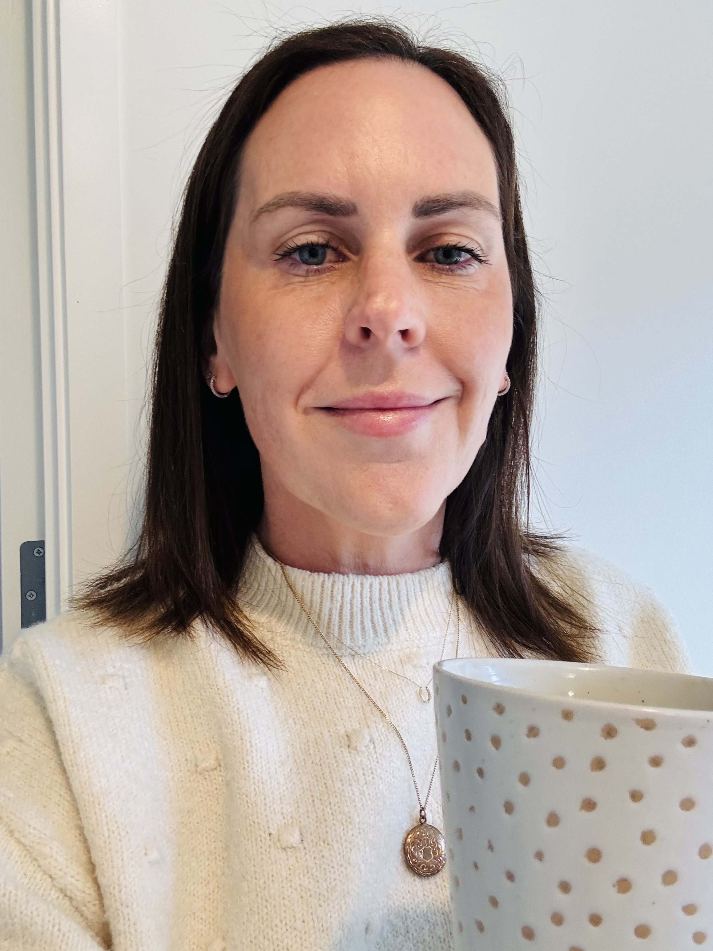 A woman with brown hair holding a white mug with gold polka dots, smiling slightly at the camera. She is wearing a cream-colored knit sweater and a necklace with a round pendant.