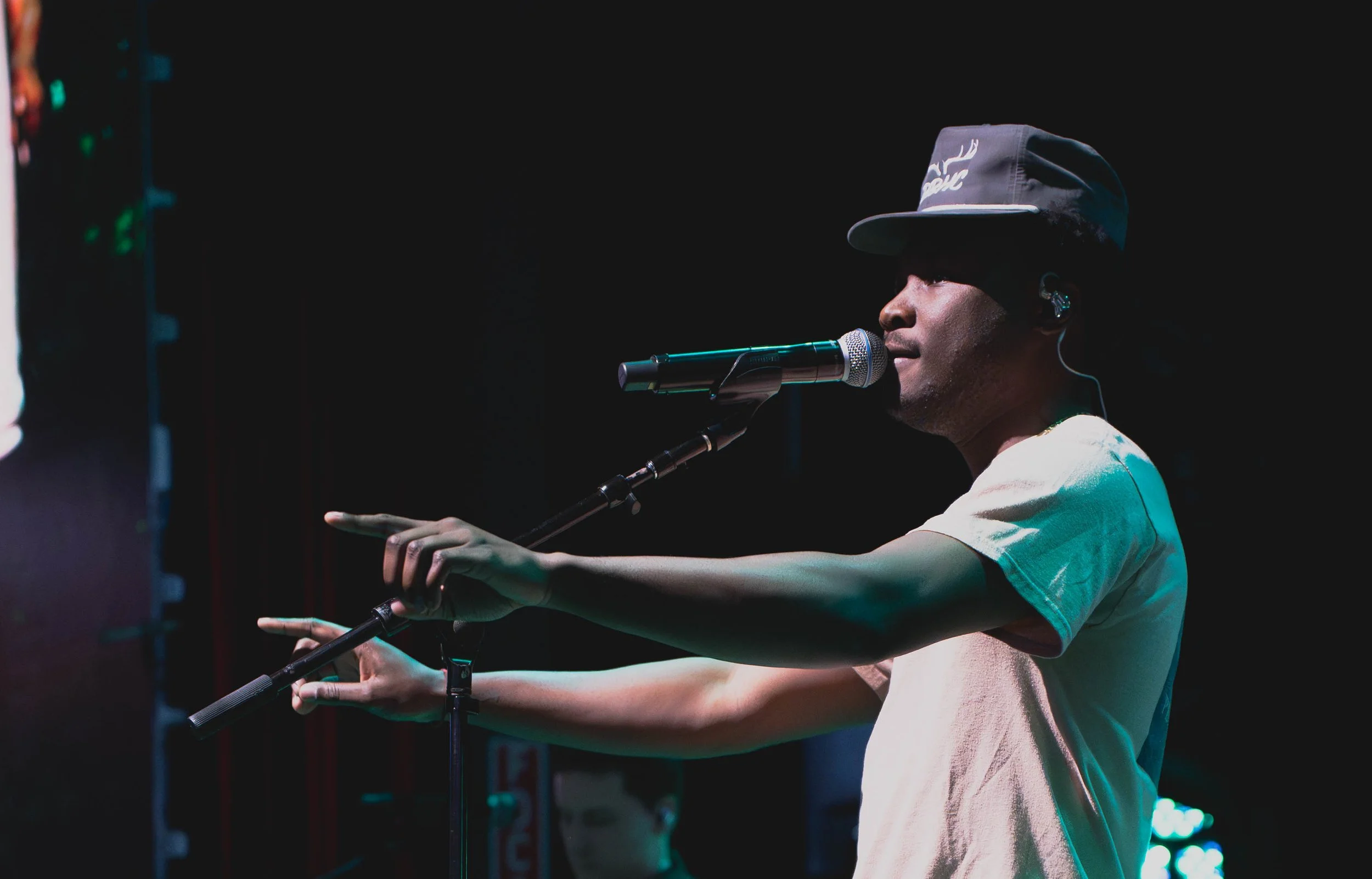 A man wearing a cap and cream-colored t-shirt performing on stage with a microphone, pointing with his hand.