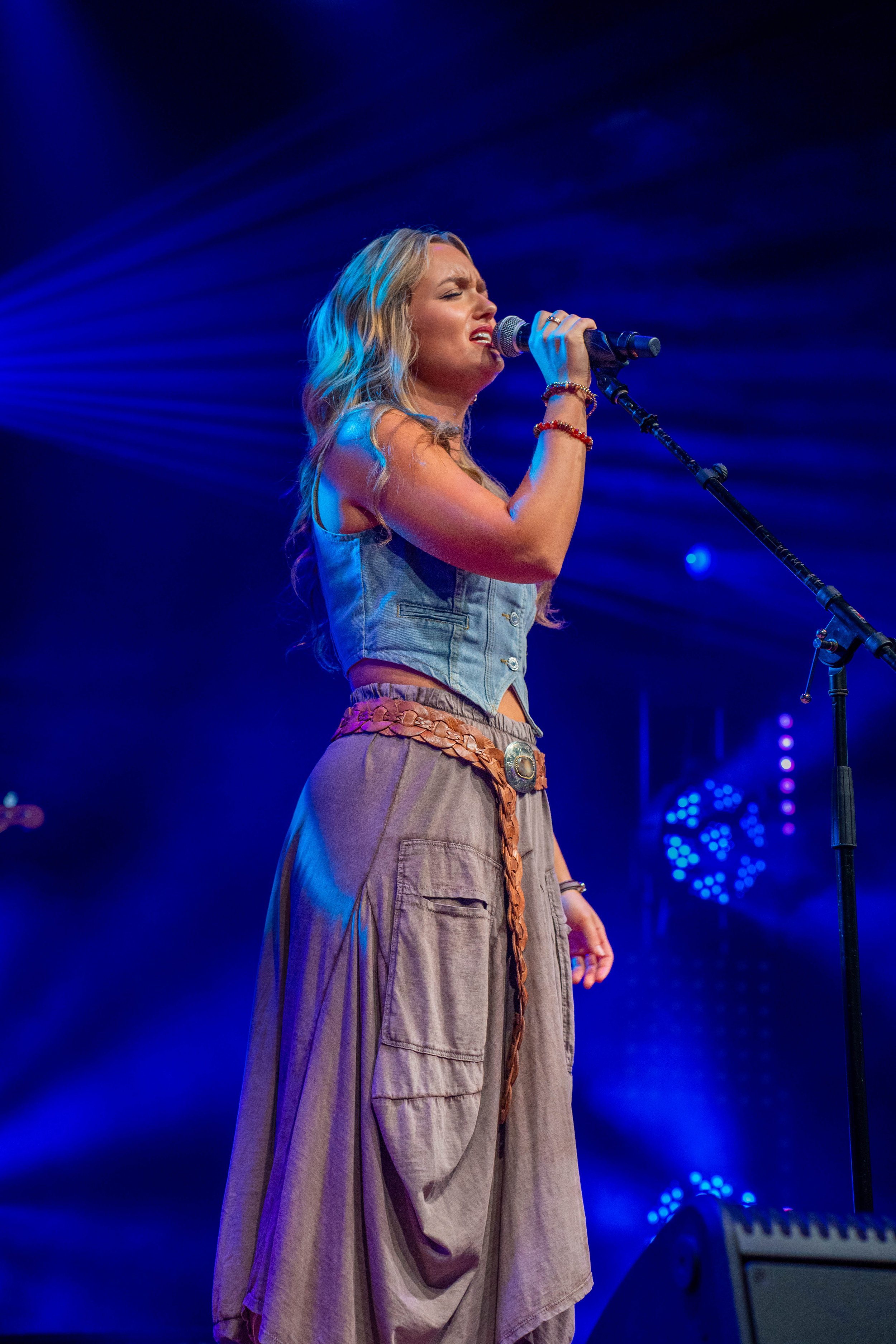 A woman singing on stage under blue lights, holding a microphone, wearing a denim vest, beige cargo pants, and a braided belt.