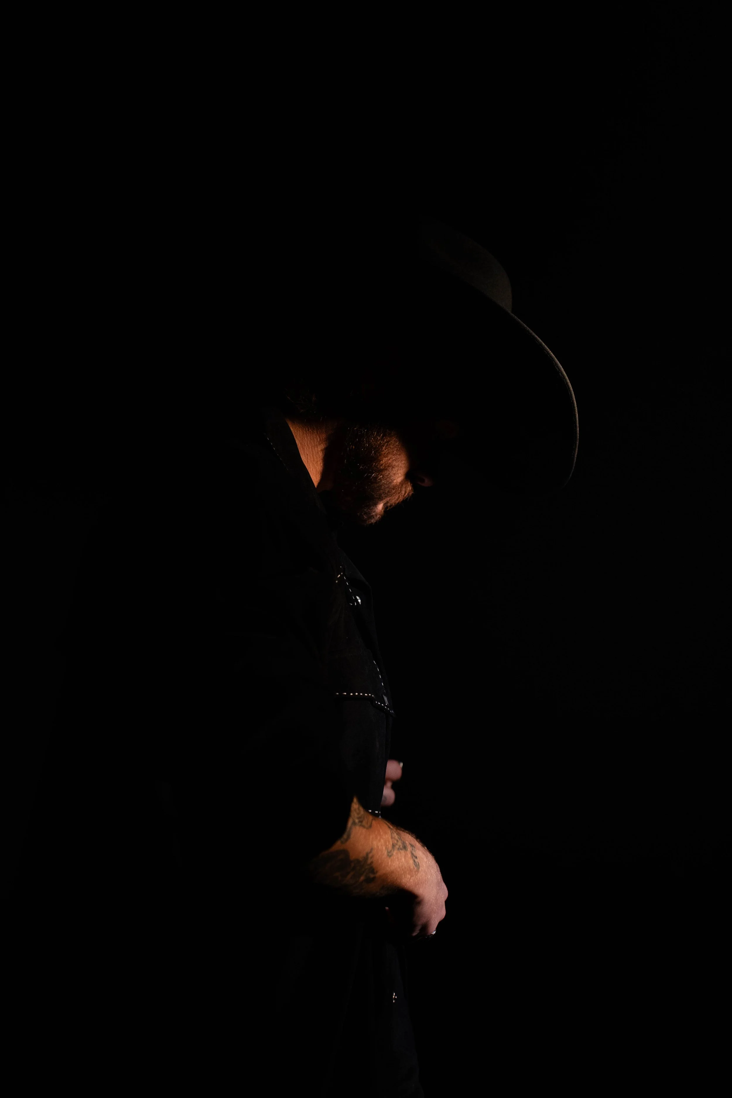 A person wearing a wide-brimmed hat, dark jacket, and tattoos on his arms, standing in a dark environment with minimal lighting highlighting his profile.