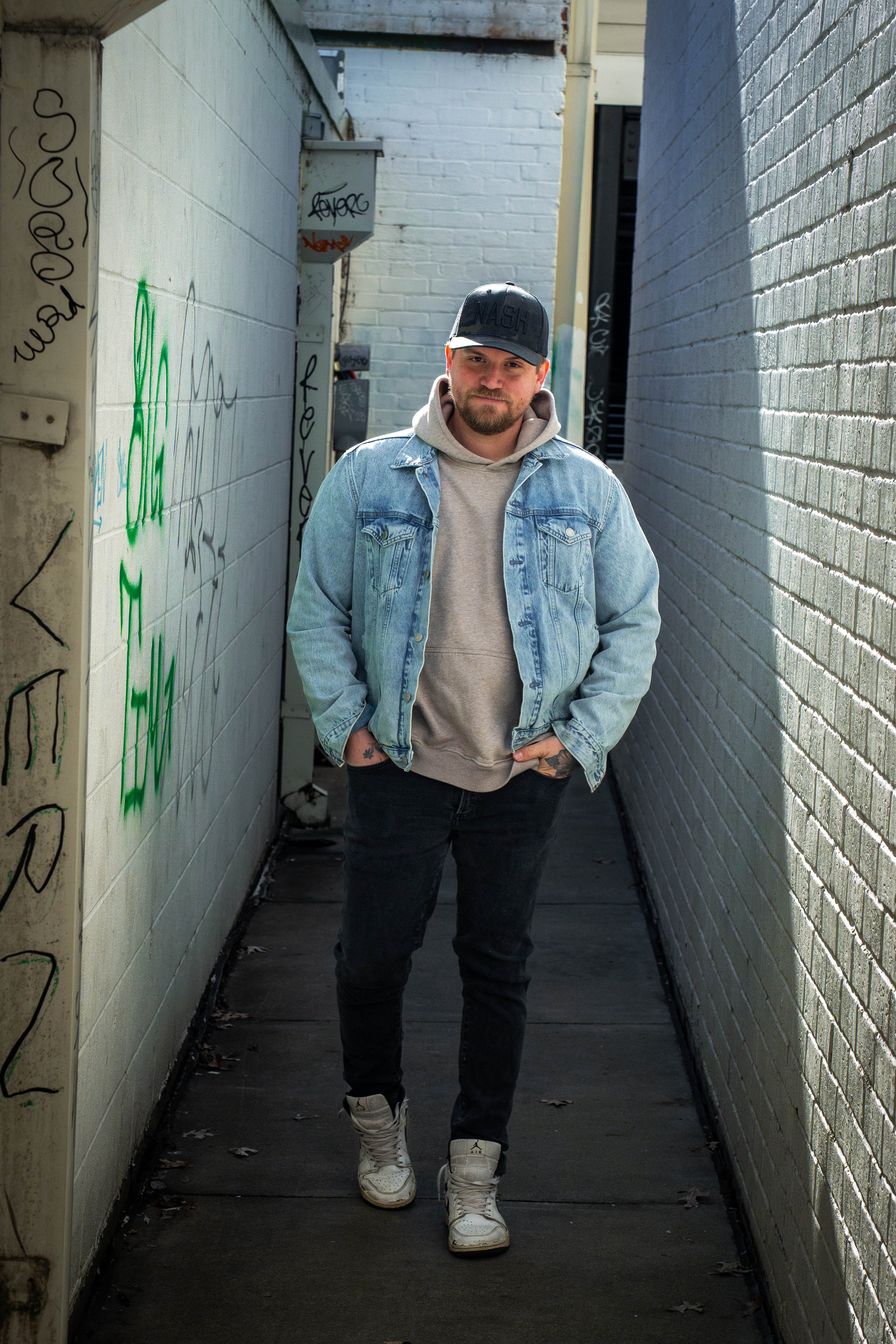 A man wearing a black cap, light gray hoodie, and denim jacket walking in a narrow alleyway with graffiti on the walls.