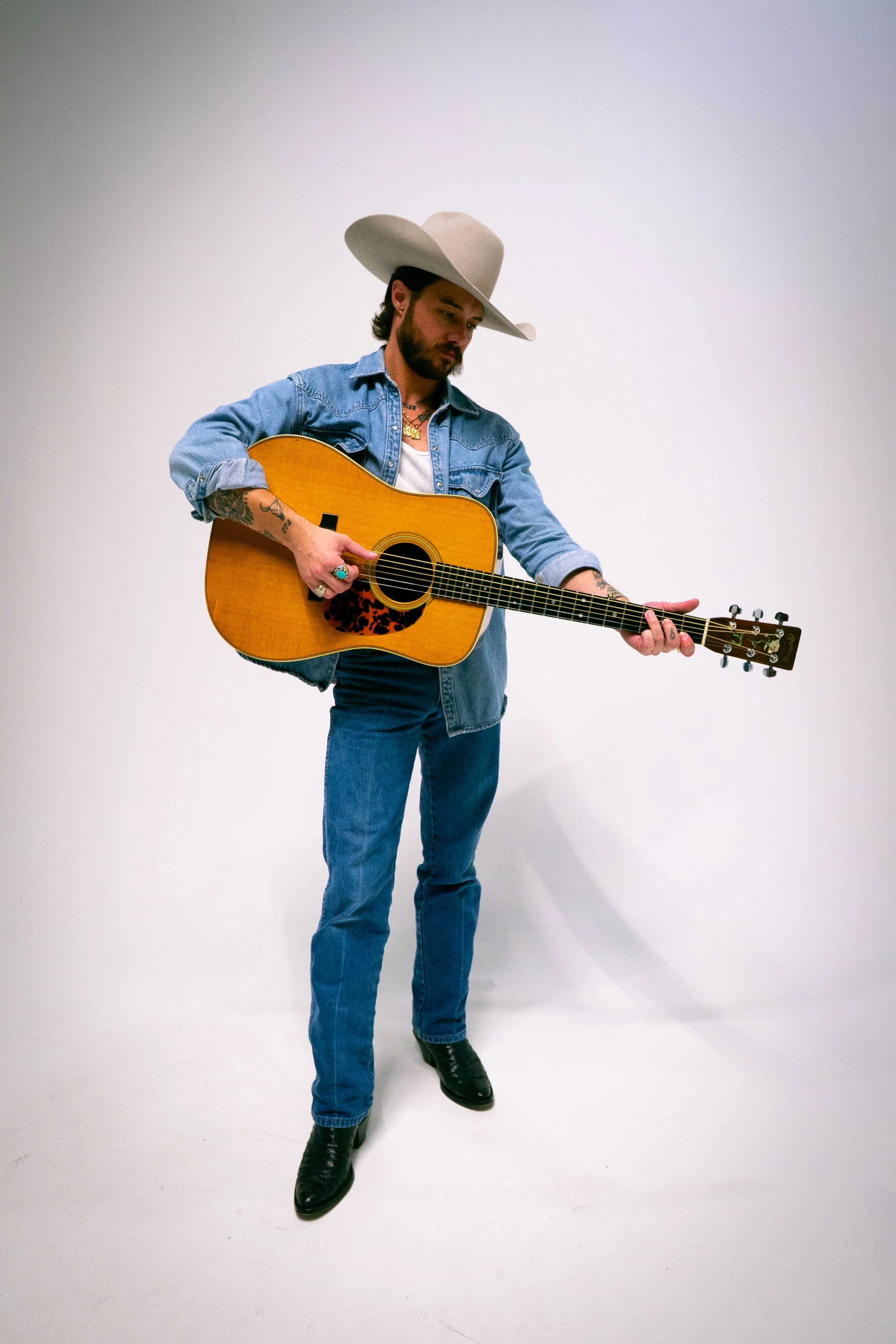 A man in a cowboy hat and denim jacket playing an acoustic guitar against a plain white background.