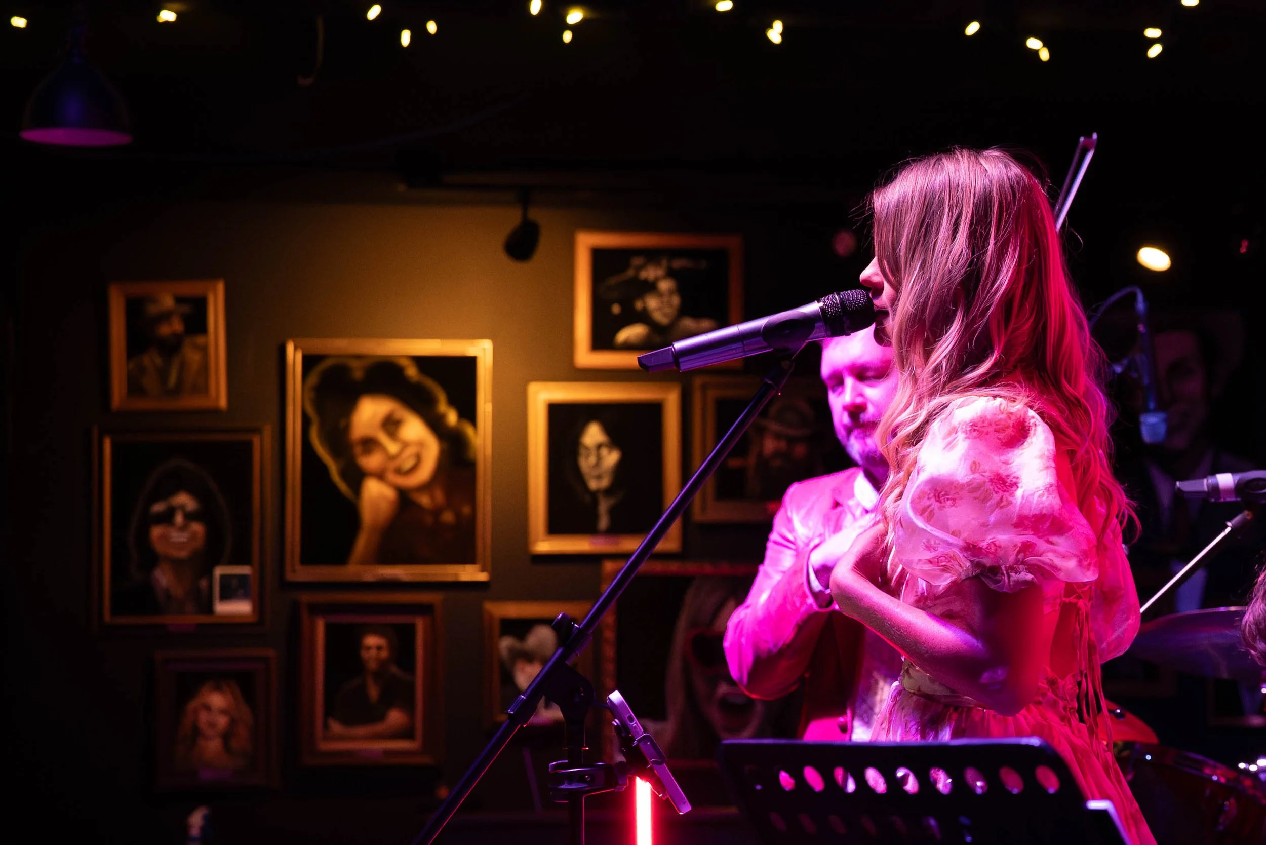 A woman with long blonde hair singing into a microphone on stage, wearing a pink floral dress. A man in a pink suit stands behind her, and there are framed portraits on the wall in the background, illuminated by pink and purple stage lights.