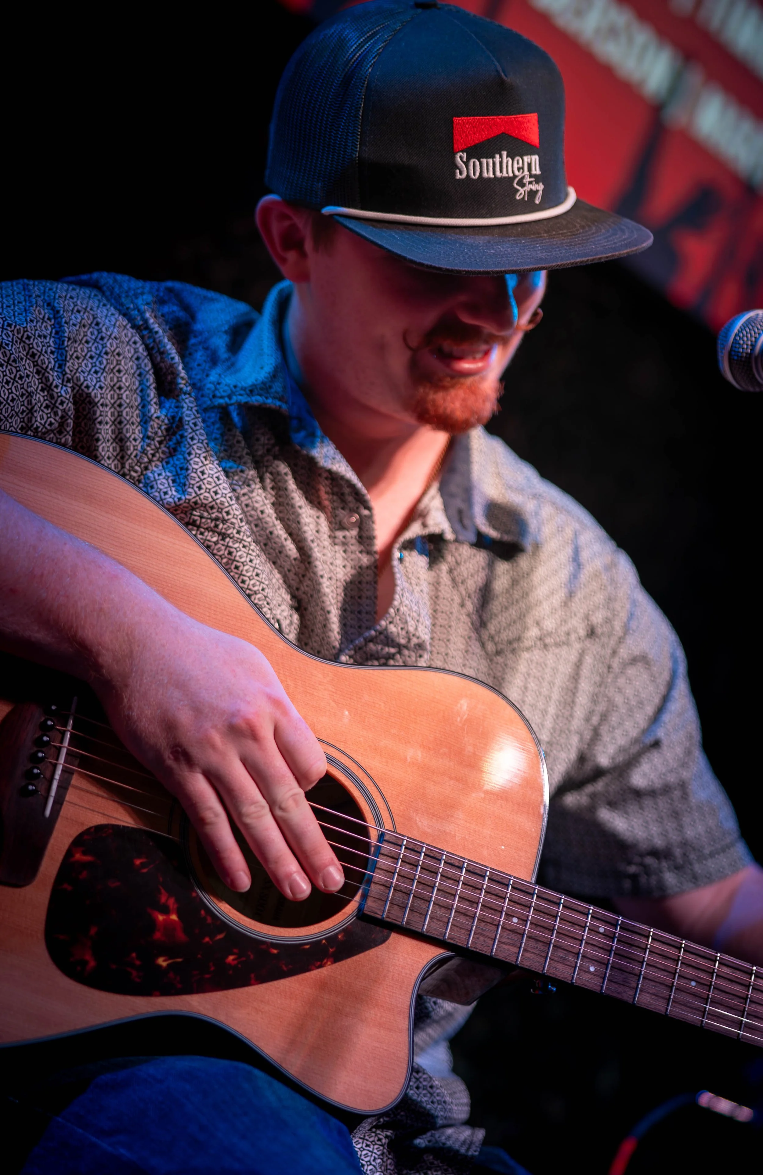 A man wearing a cap that says 'Southern' is playing an acoustic guitar and singing into a microphone on stage.
