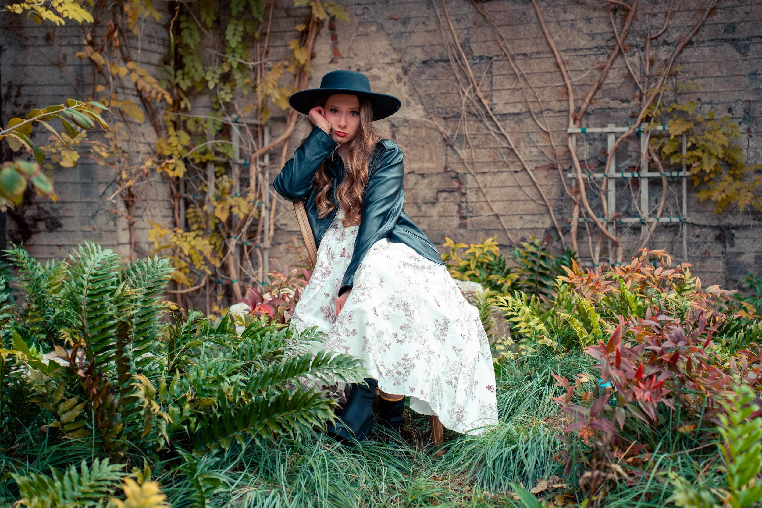 Young woman sitting in garden surrounded by green and red plants, wearing a black wide-brimmed hat, a shiny black jacket, and a long floral dress, with a distressed brick wall in the background.