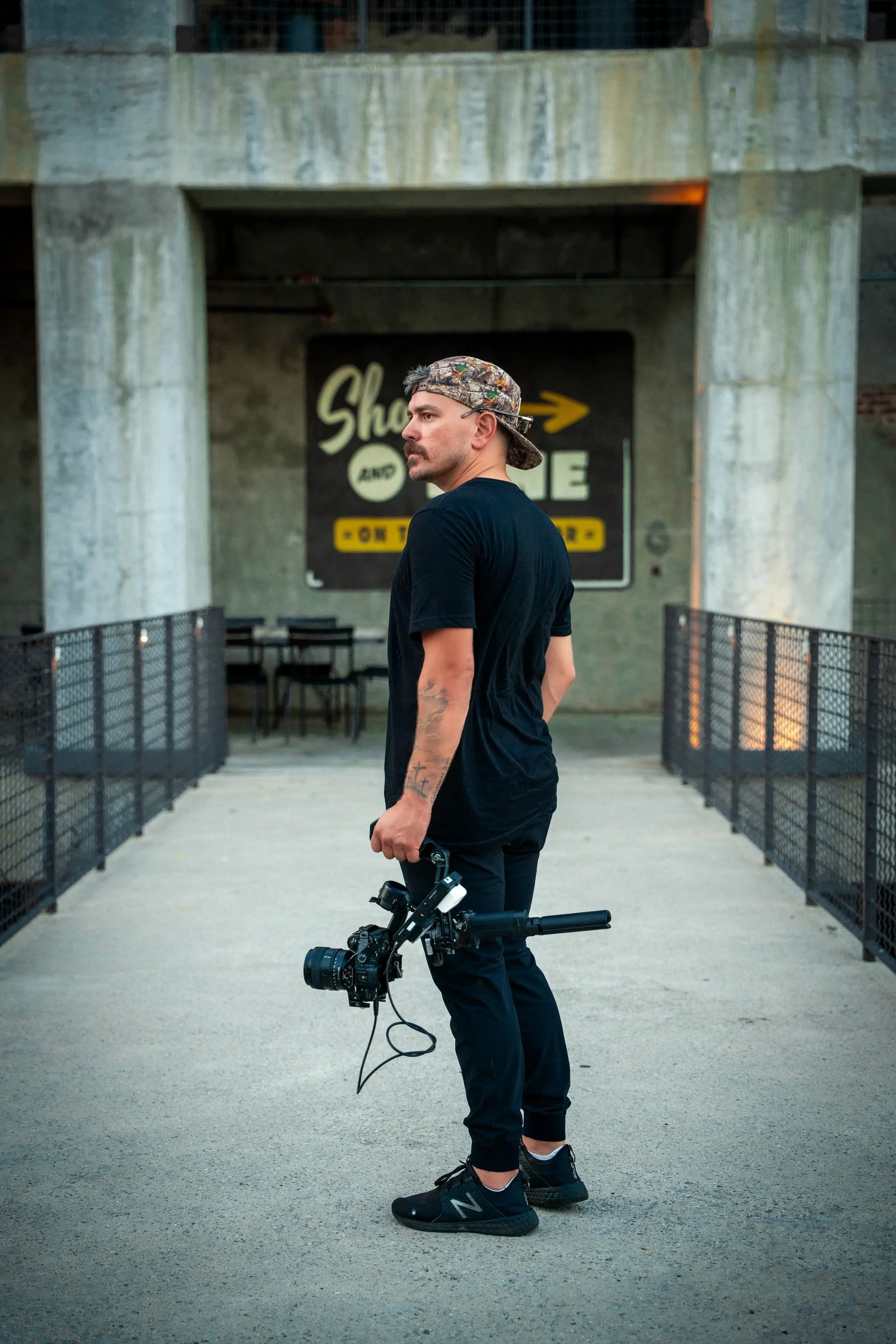 A young man with tattoos on his left arm, wearing a camouflage baseball cap backwards, black t-shirt, black pants, and black sneakers, stands on a concrete walkway holding a camera in his right hand. The background features concrete pillars and a sign that reads "Shoot and Share on the Bridge" with an arrow pointing to the right.