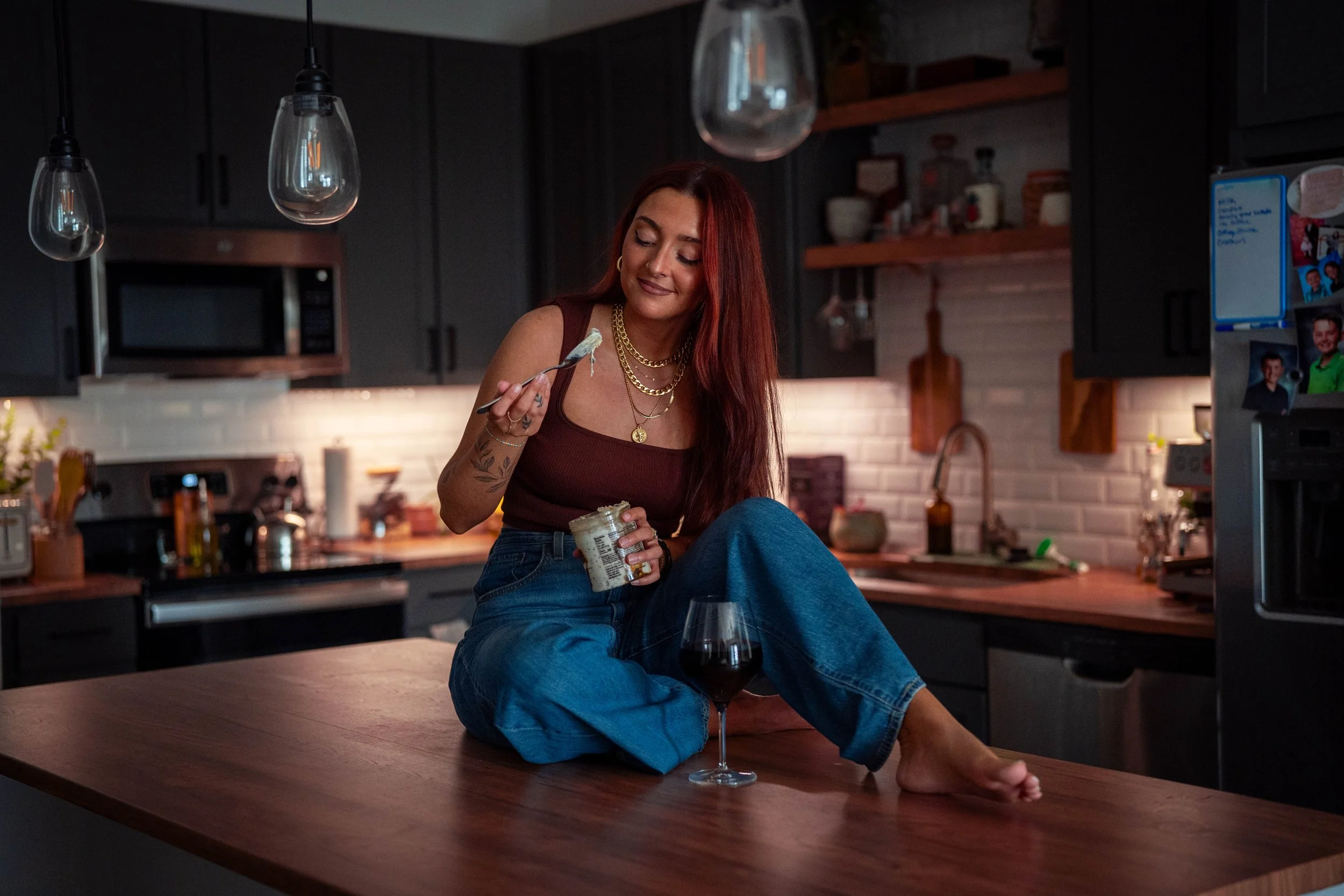 A woman with long red hair and tattoos on her arm sitting on a wooden kitchen counter, eating from a jar with a spoon, with a glass of red wine nearby. The kitchen has dark cabinets, a microwave, pendant lights, and a white brick backsplash.