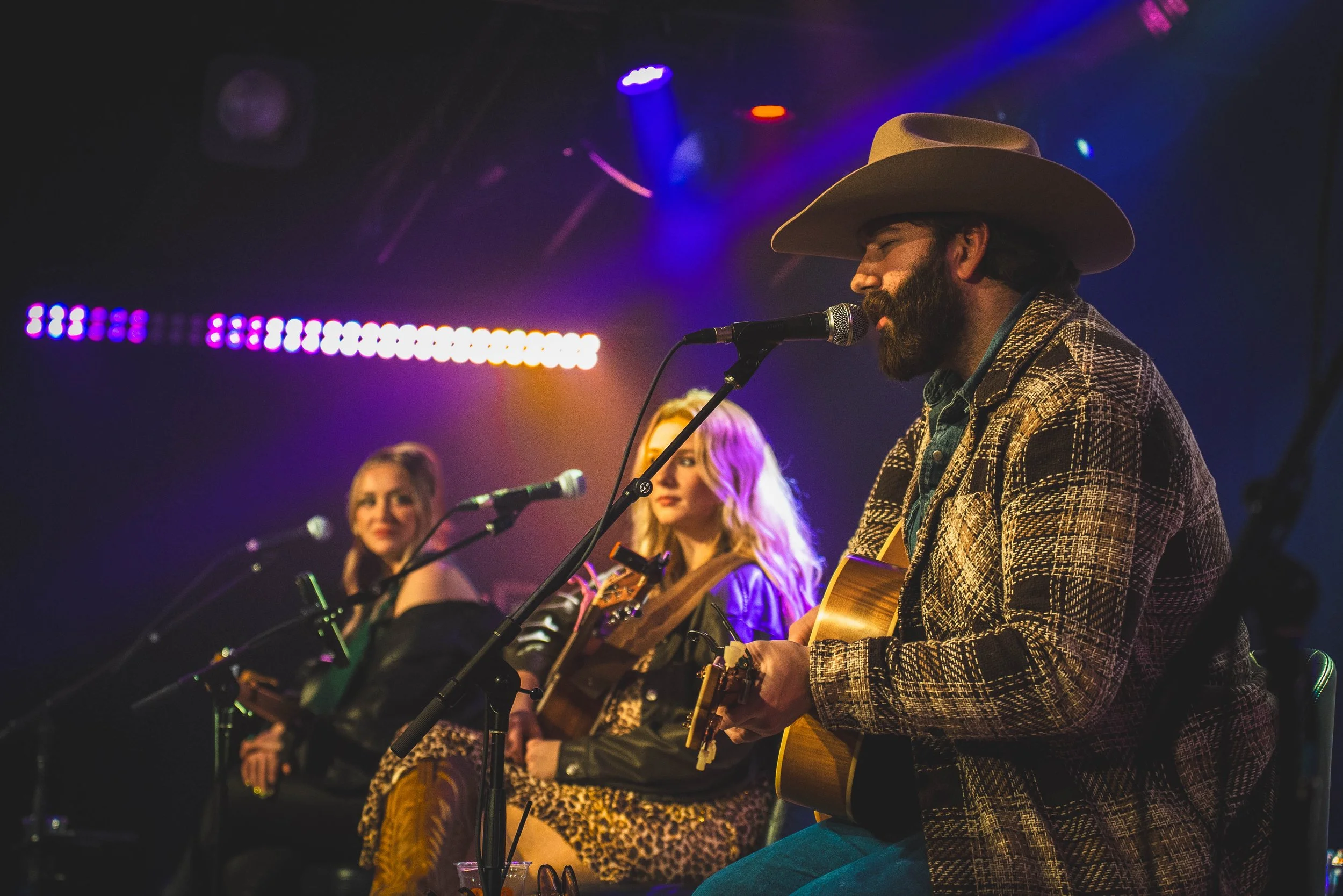 A man with a beard wearing a cowboy hat and a patterned jacket playing acoustic guitar into a microphone, with two women sitting beside him, one with blonde hair and the other with blonde hair in waves, both also with microphones on stage with colorful stage lighting.