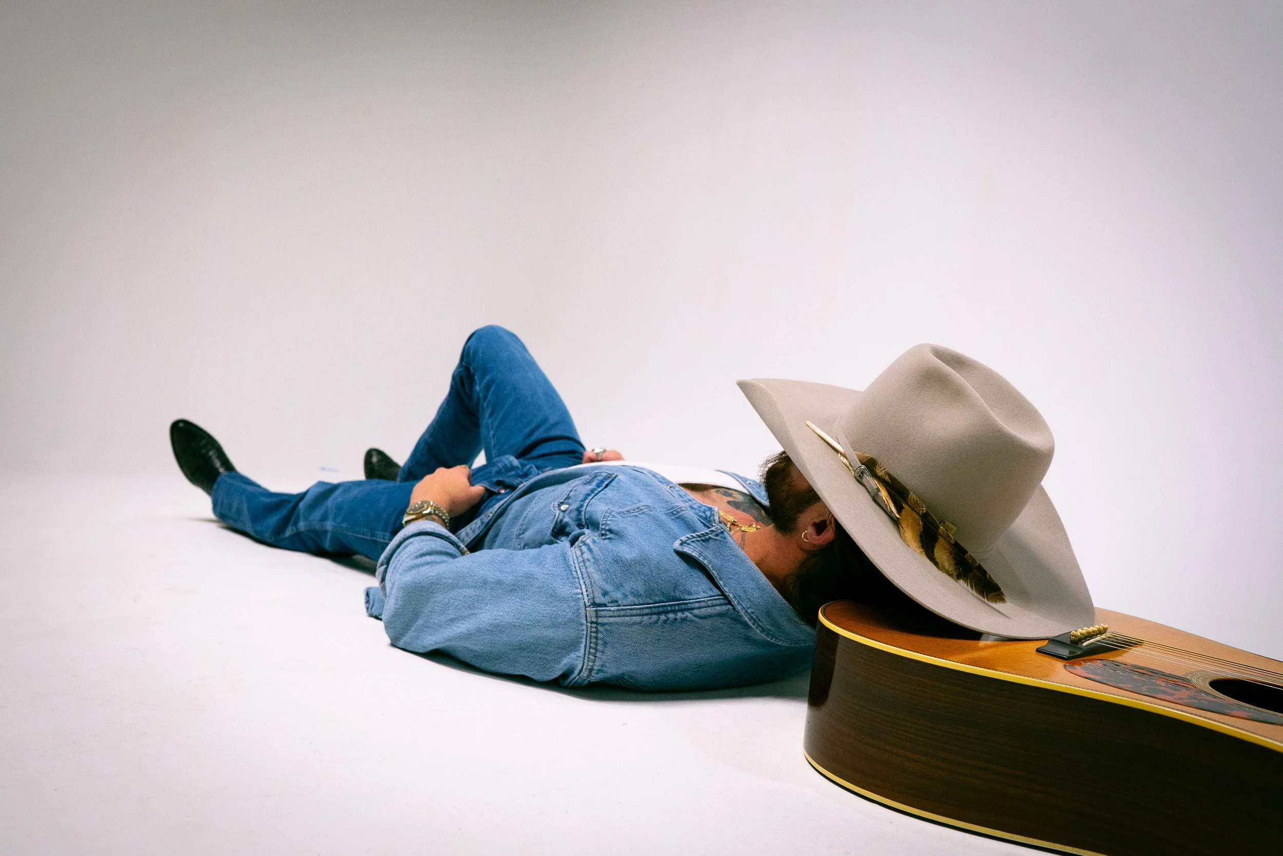 Person lying on the floor with their head resting on a large guitar, wearing a wide-brimmed hat, denim jacket, and jeans against a plain white background.