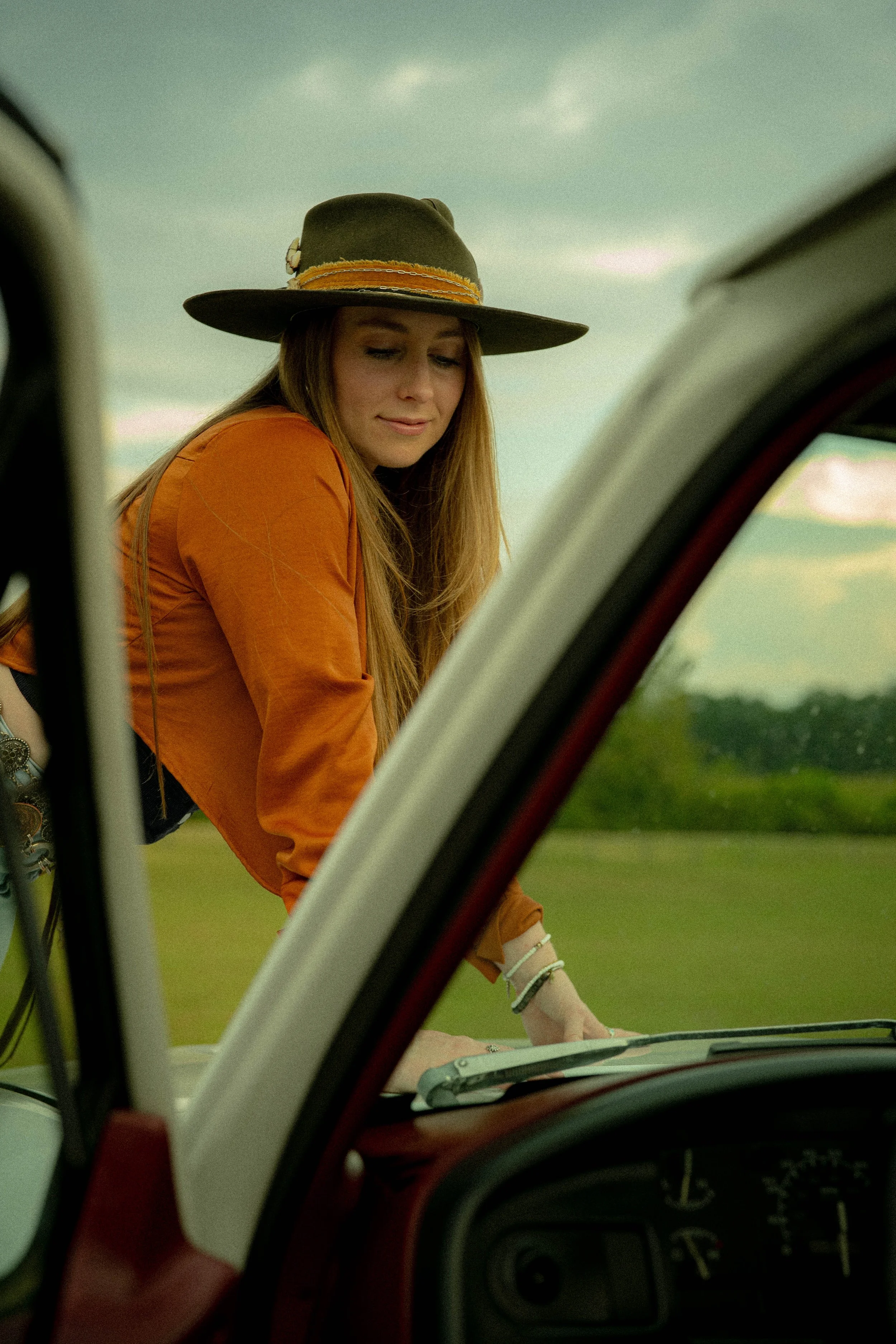 A woman with long hair wearing a wide-brimmed hat and an orange jacket, leaning over a vintage vehicle outdoors with a grassy field and cloudy sky in the background.