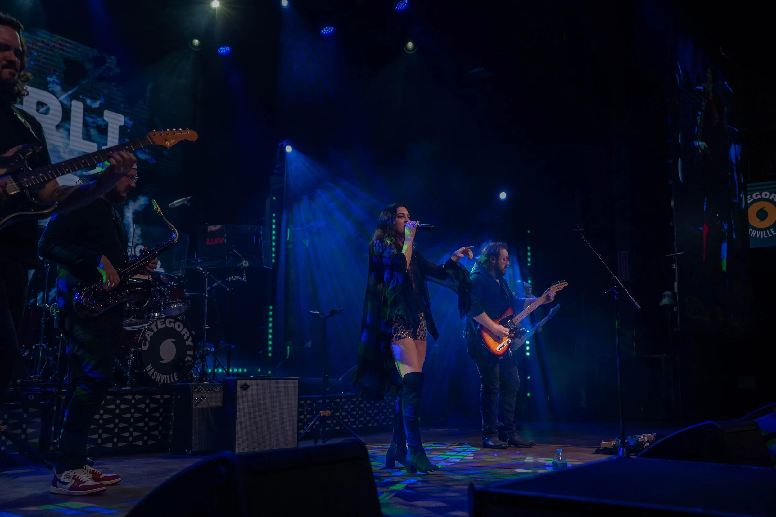 A live concert performance with a female singer and four male guitarists/band members on stage, illuminated by blue and purple stage lighting, with a backdrop displaying the words 'CATEGOR' and 'NASHVILLE'.