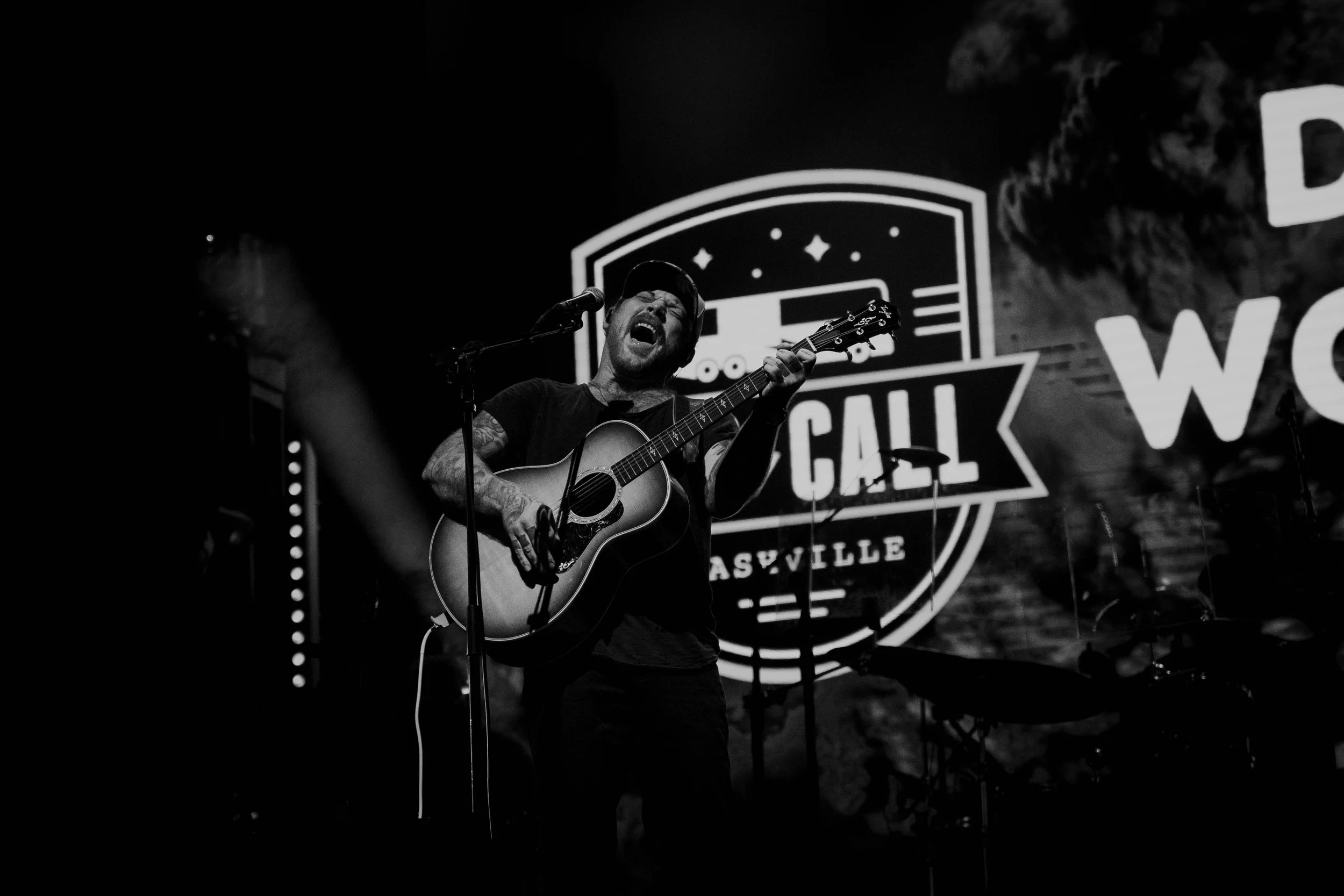A male musician passionately singing and playing an acoustic guitar on stage with Nashville's Bluebird Cafe logo in the background, in black and white.