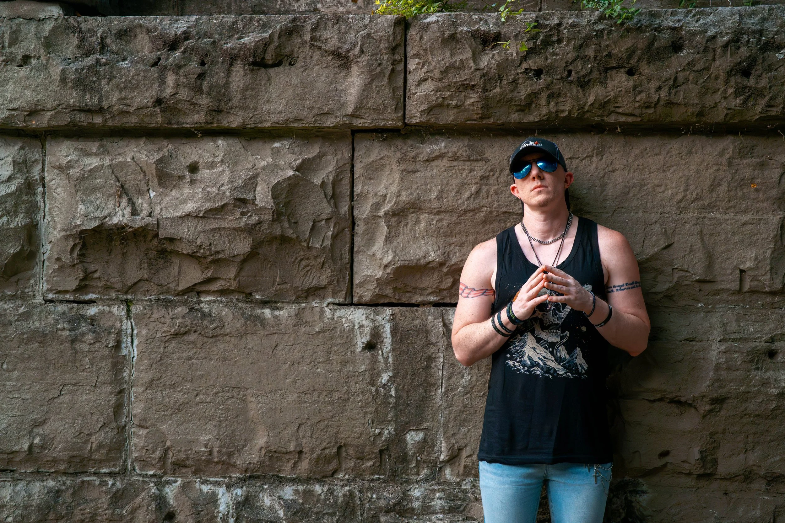 A young man with tattoos, wearing a black tank top with a skull and mountain design, sunglasses, a black cap, and multiple wristbands, standing against a stone wall with his hands clasped together.