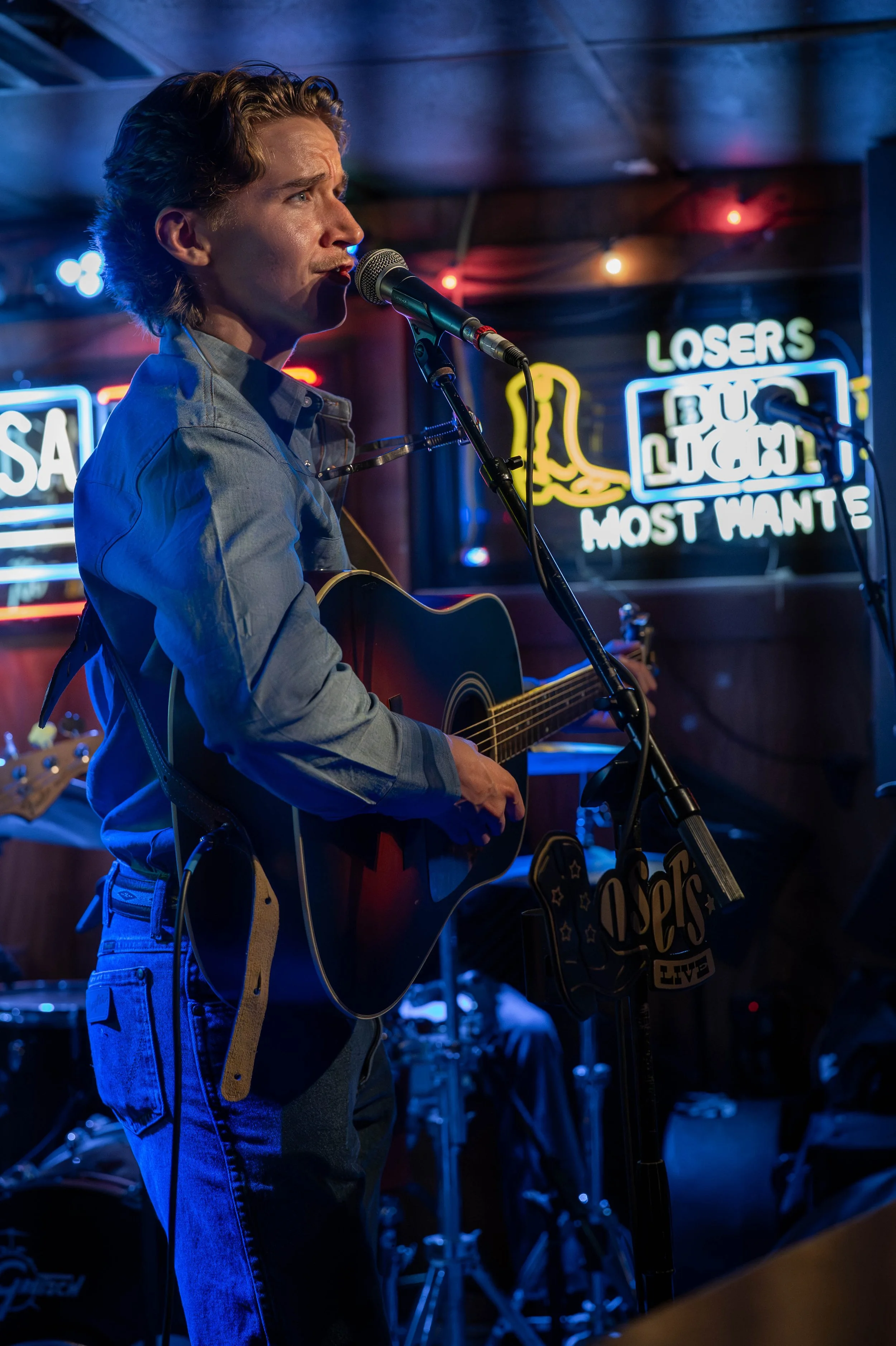 A man playing guitar and singing into a microphone in a dimly lit bar with neon signs in the background.