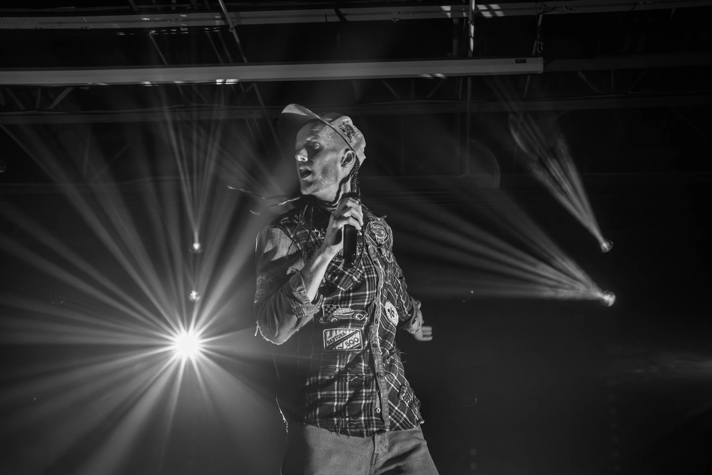 A male performer on stage holding a microphone, with dramatic lighting and spotlights creating rays of light in the background, in black and white.