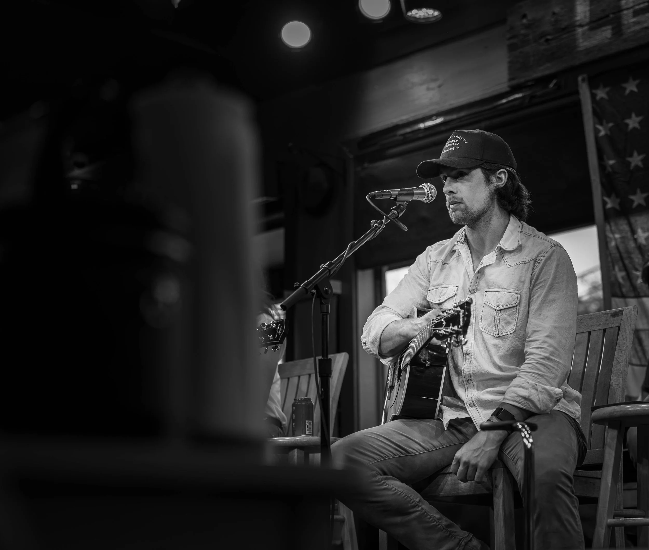 A man playing an acoustic guitar and singing into a microphone on a stage, with an American flag in the background, in a black-and-white photo.