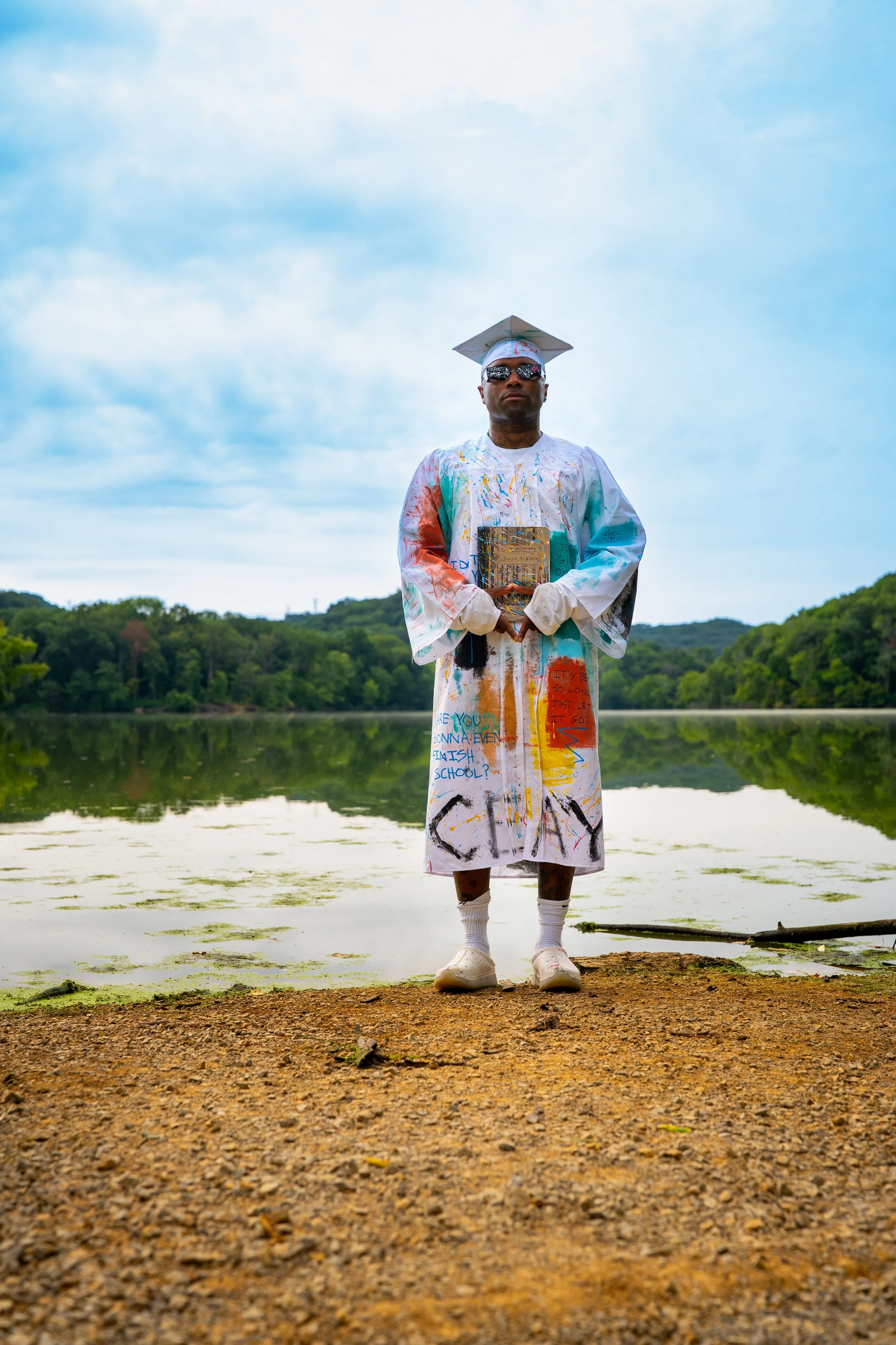 A man stands on a dirt path by a lake, wearing a white graduation gown covered in colorful paint, holding books, with a graduation cap and sunglasses, surrounded by green trees and a cloudy sky.