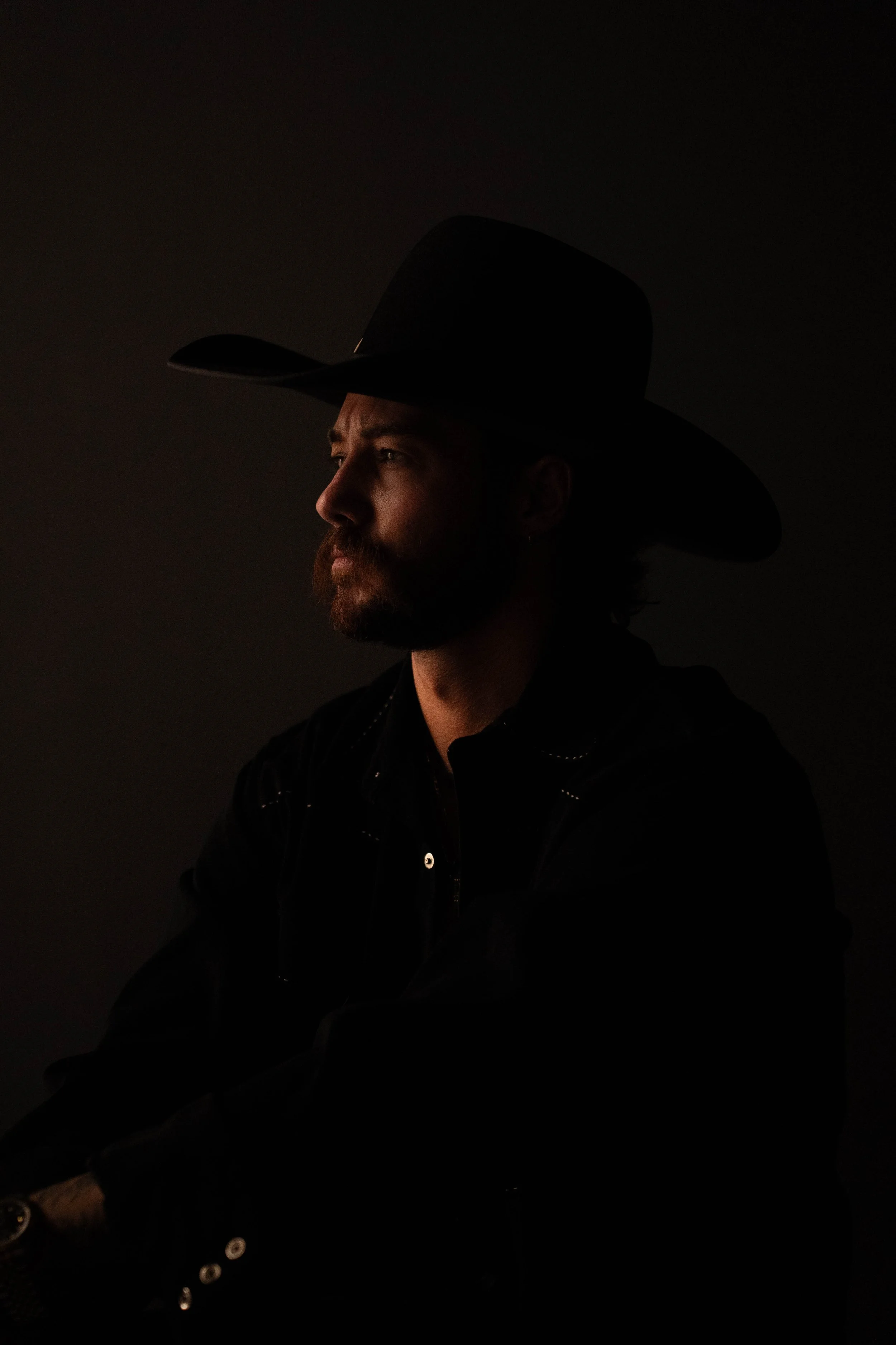 A man wearing a black cowboy hat and black shirt, looking to the left with a serious expression, against a dark background.