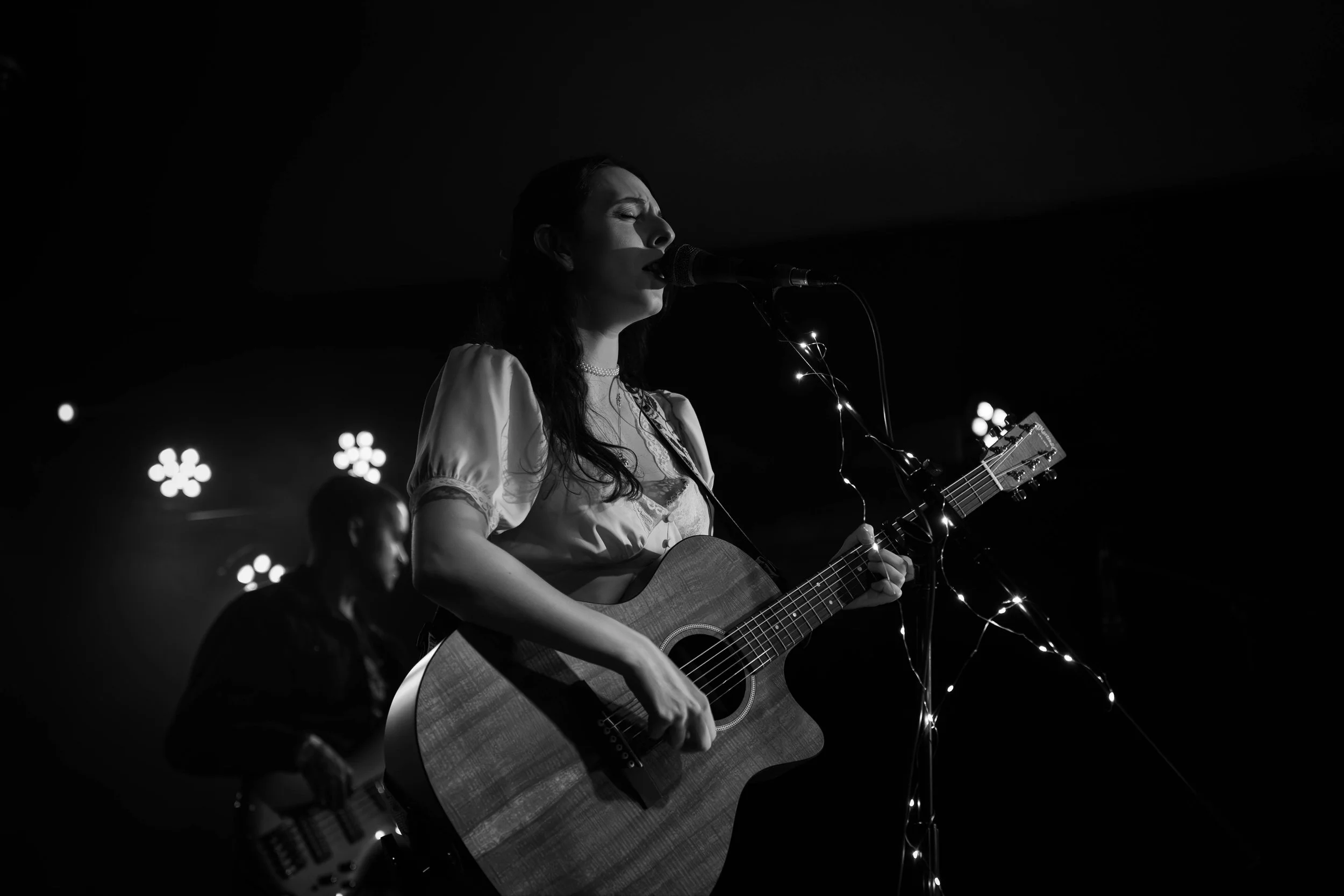 Black and white photo of a female musician singing into a microphone and playing an acoustic guitar, with a male musician in the background playing an electric bass guitar, illuminated by stage lights.