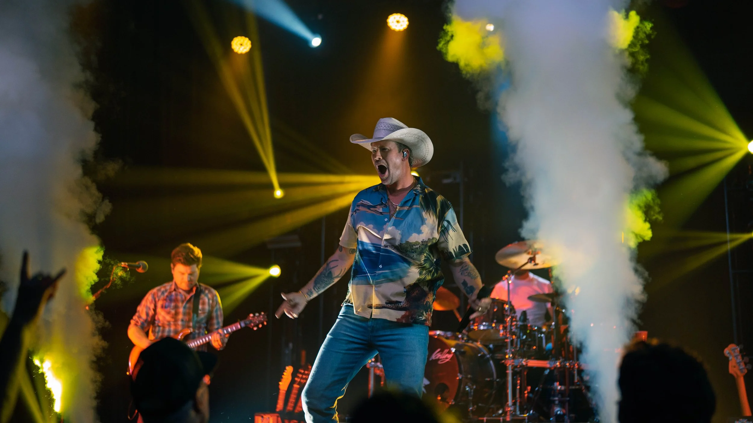 A male country singer wearing a cowboy hat, colorful Hawaiian shirt, and jeans performing on stage with band members, bright yellow stage lights, and smoke effects.