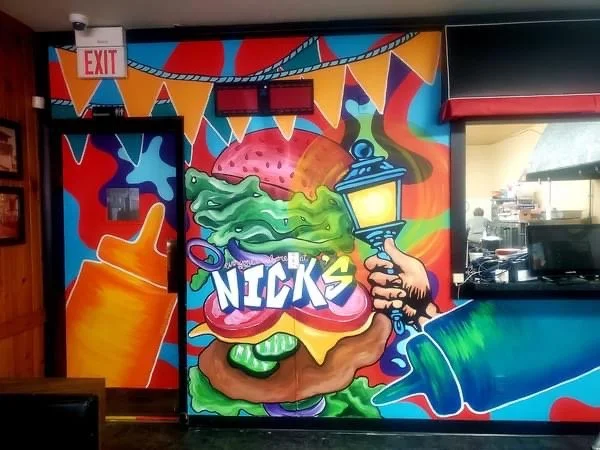 Colorful mural showing a large burger with lettuce, tomato, cheese, and a pink bun, holding the text 'Nick's.' A hand holding a lantern shines light onto the burger, with abstract shapes and a rainbow of colors in the background.