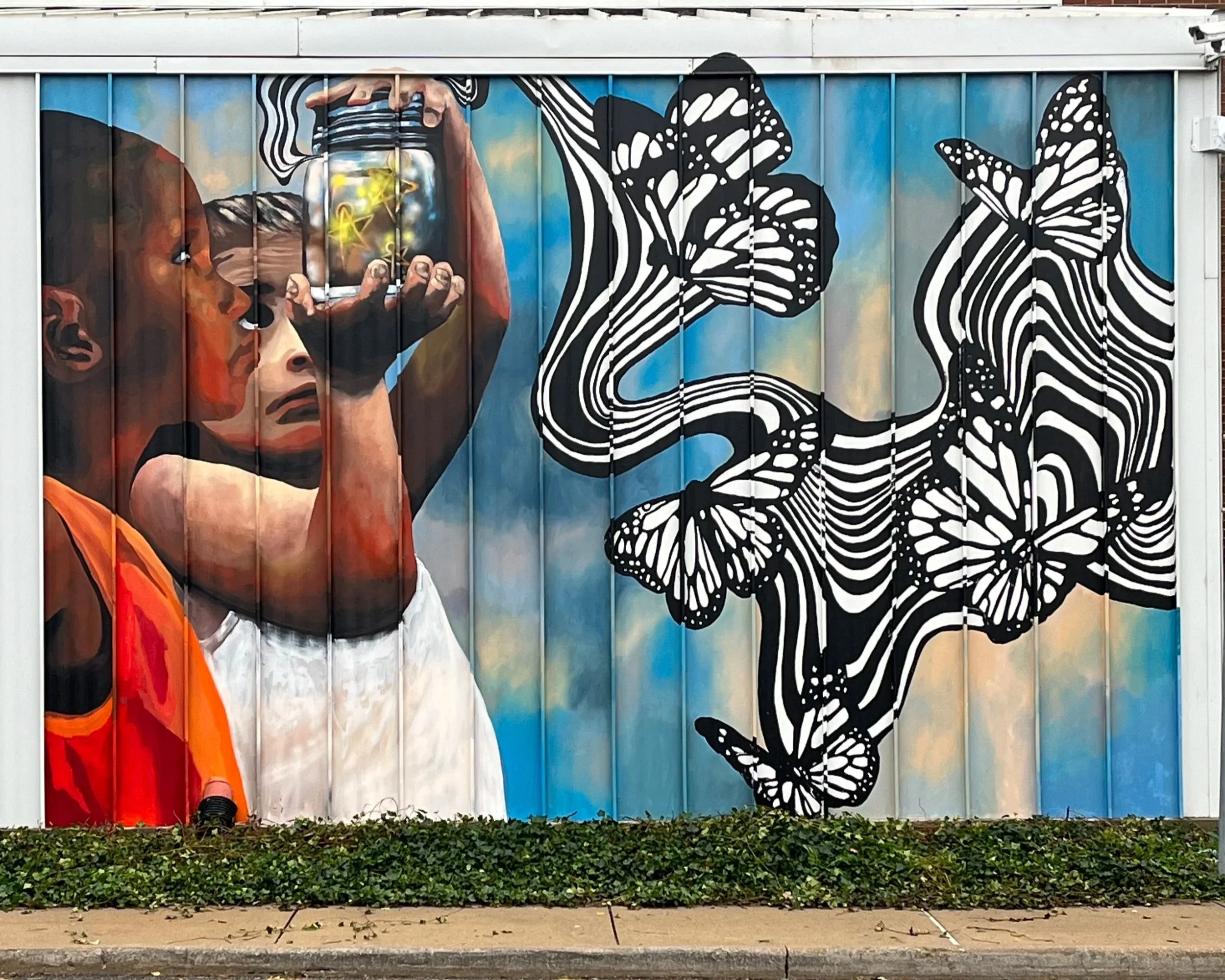 Street art mural portraying a boy looking through a jar with bright yellow lighting inside, surrounded by black and white patterned butterflies, against a partly cloudy blue sky background.