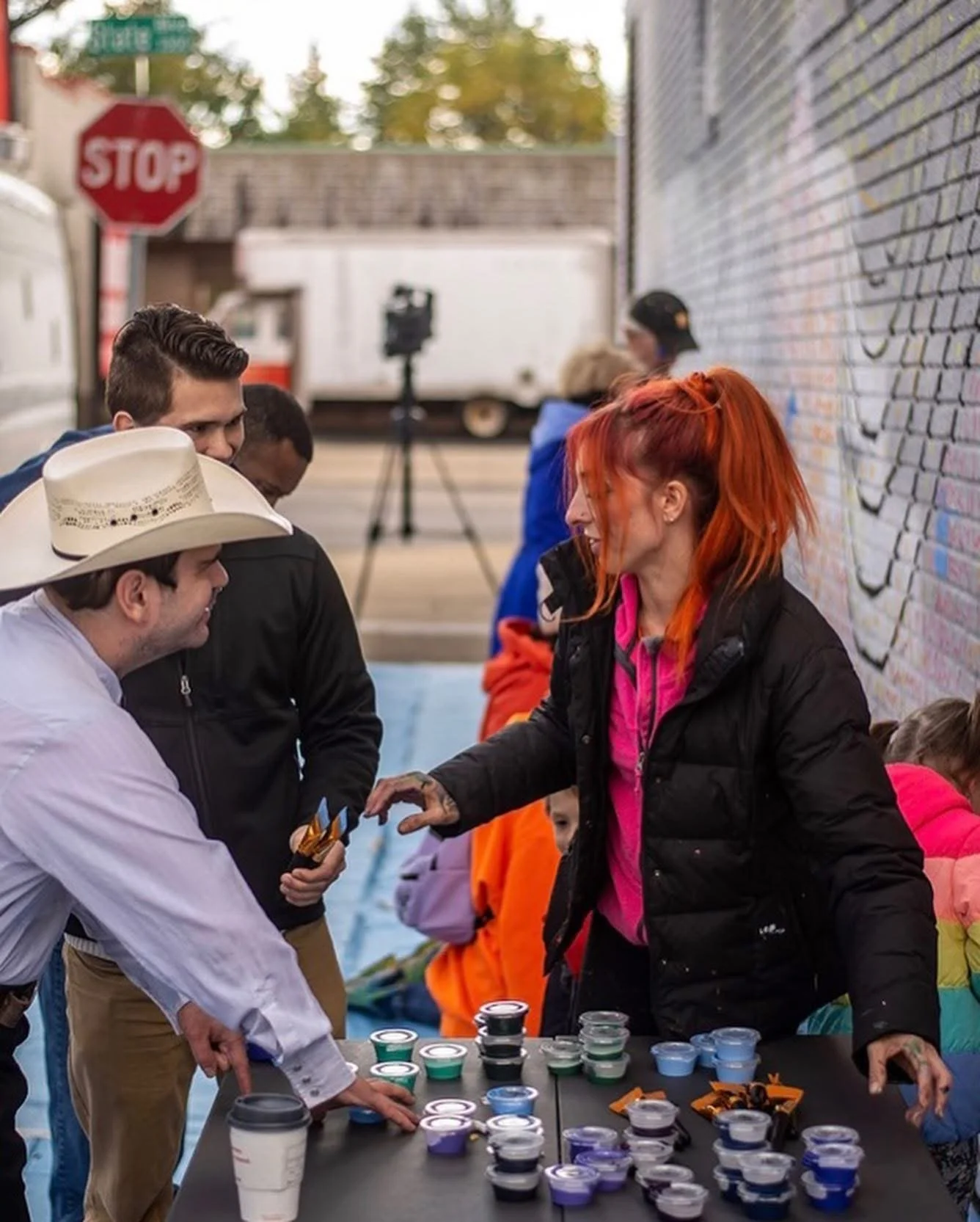 A woman with orange hair wearing a black jacket is giving small containers of colorful paint to a man with a white hat and white shirt at an outdoor event. Several people are in the background, along with a table filled with small paint containers, cups, and medals. There is a stop sign and street signs visible, indicating a city street setting.