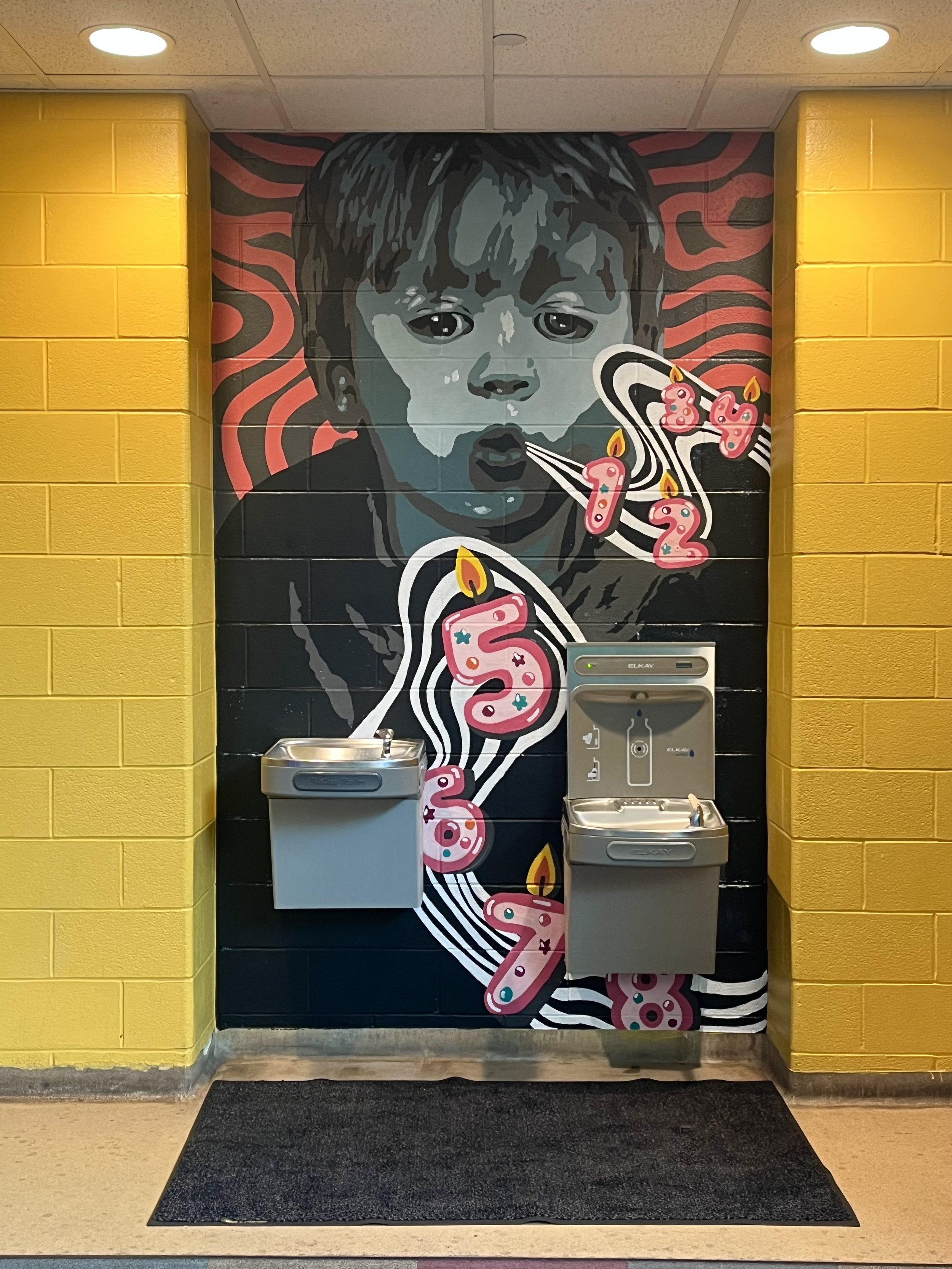 A mural of a child's face blowing out birthday candles on cupcakes, with a yellow brick wall on either side and water fountains in front.